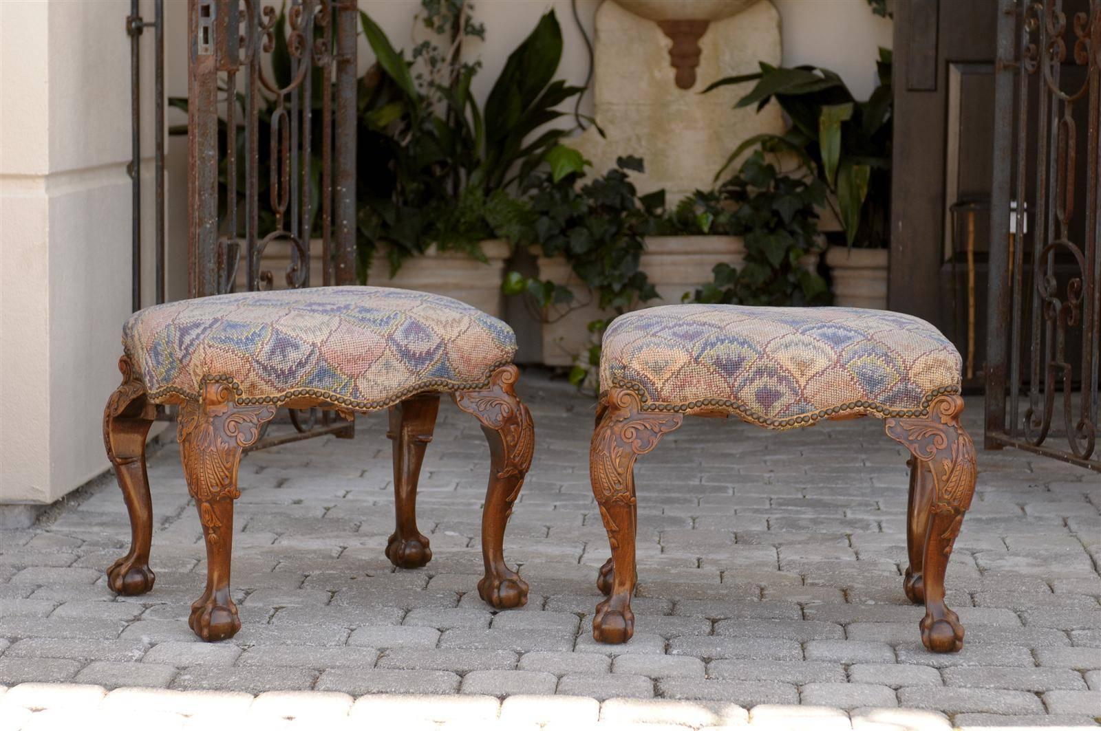This pair of English Early Georgian style wooden stools from the mid 19th century features rectangular needlepoint upholstered seats with blue, pink and purple tones and nailhead trim. The stools are raised on exquisite carved wood cabriole legs,