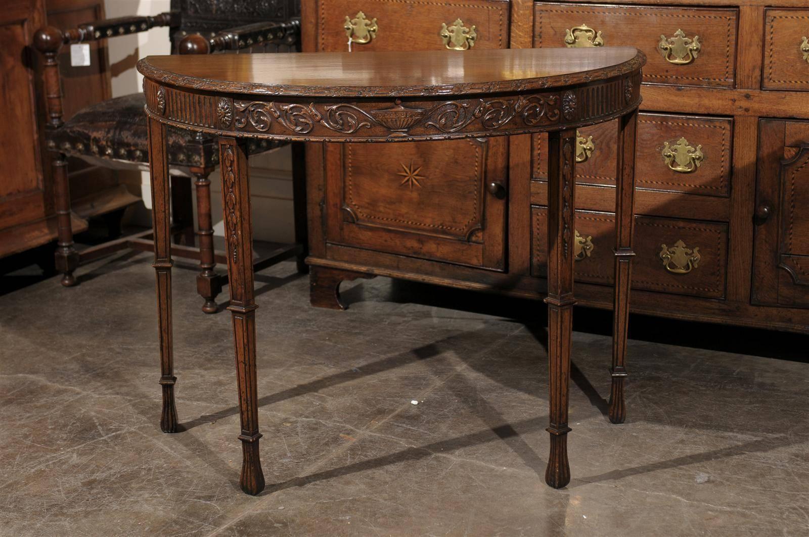 This English wooden demi-lune table from the turn of the century (19th to 20th) features a half-moon shaped top with a carved and beveled edge. The skirt is delicately carved with a cassolette in the center, flanked with rinceaux friezes on each