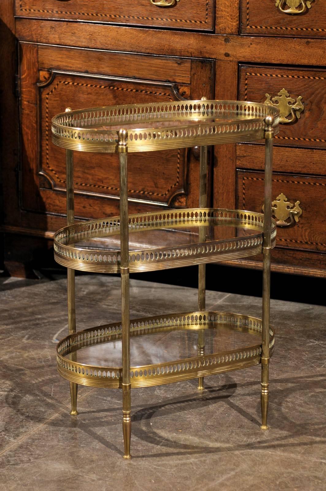 A petite French brass oval table with glass inset shelves from the mid-20th century. This French brass table inspired by the Maison Jansen style features three tiers of clear glass shelves decorated with an oval pierced gallery edging. This small