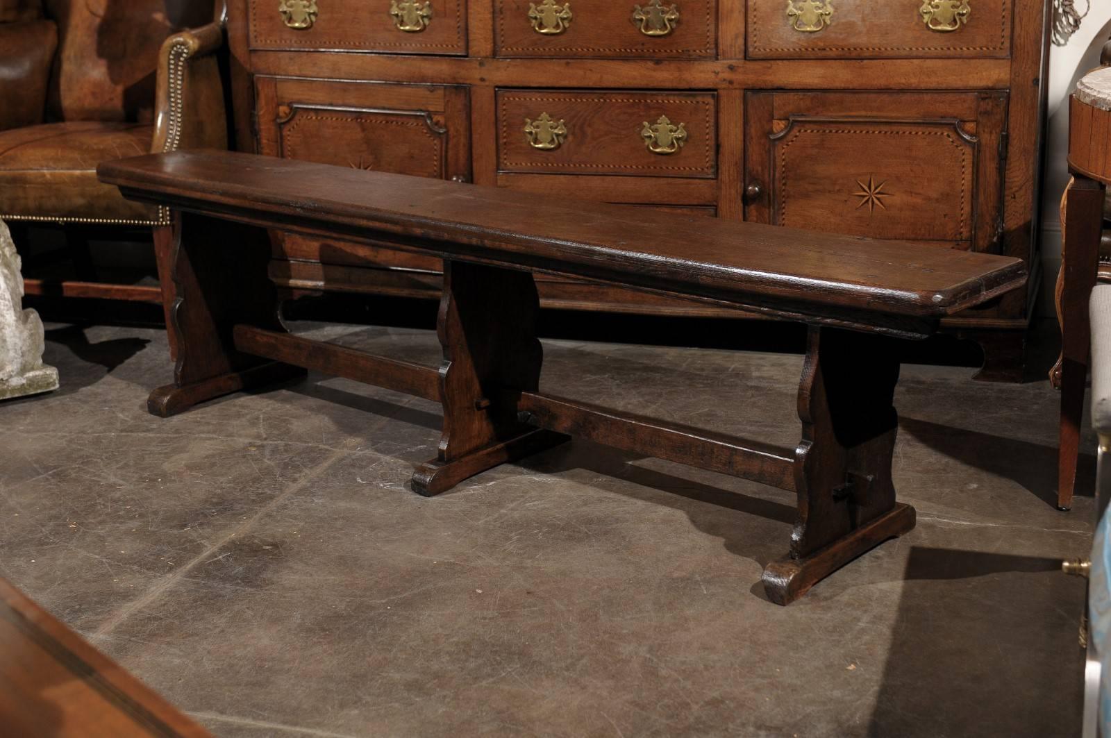 A 19th century long wooden bench with a rectangular molded seat over three trestle crossbow-shaped supports, joined by a straight stretcher protruding from the outer legs, offering numerous convivial sitting places. The rich and dark patina along