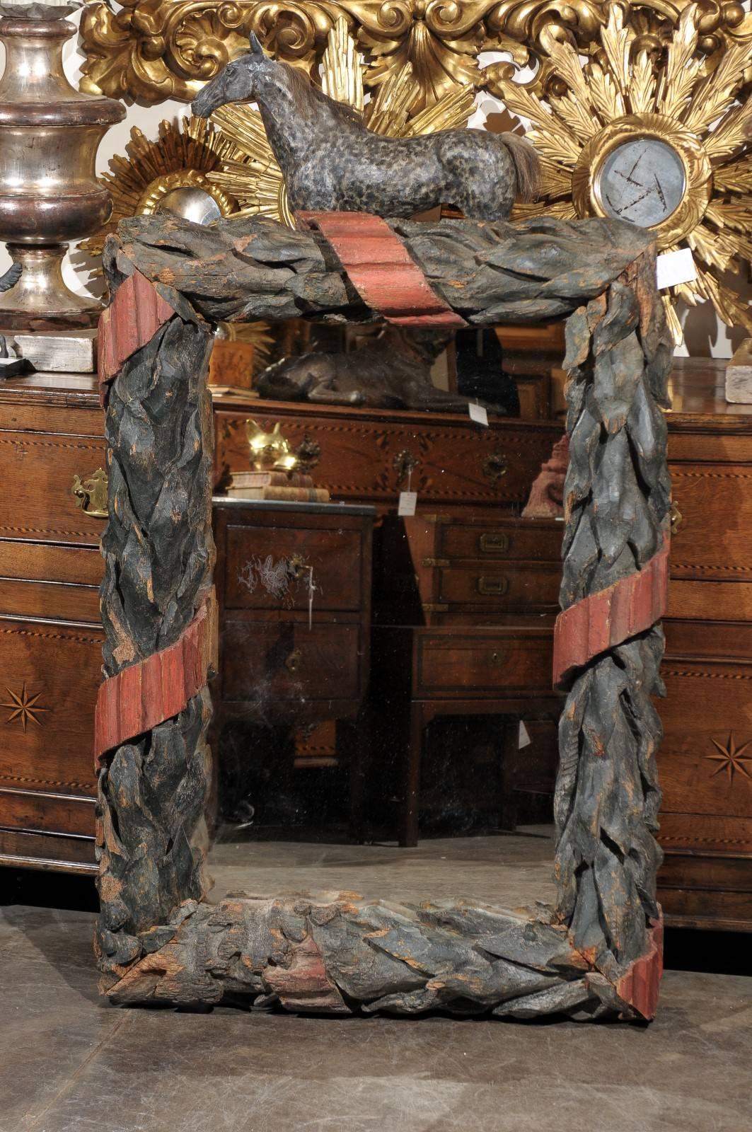 This large Black Forest wood mirror features a frame of carved green-painted oak leaves arranged in a rectangular wreath and tied together by a twirling red-painted reeded ribbon reminiscent of the antique fasces. The distressed paint gives