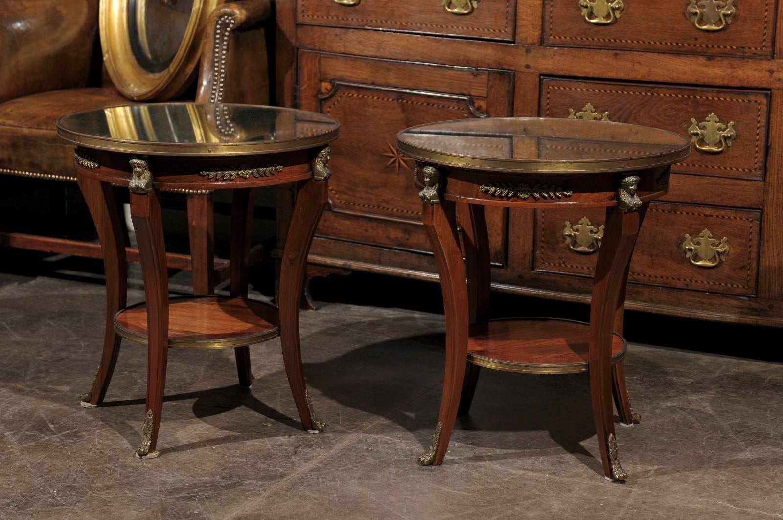 A pair of French Empire style accent tables from the early 20th century. Each table features a circular top of round shape set into a brass frame. The aprons are decorated with bronze motifs, typical of the Empire style, accentuated by the presence