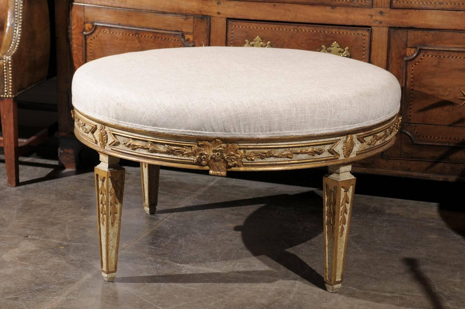 This Italian early 20th century round painted wood ottoman features a muslin upholstered top over a beautifully carved skirt. The painted wooden body is gracefully adorned with giltwood motifs made of foliage friezes marked in their center with