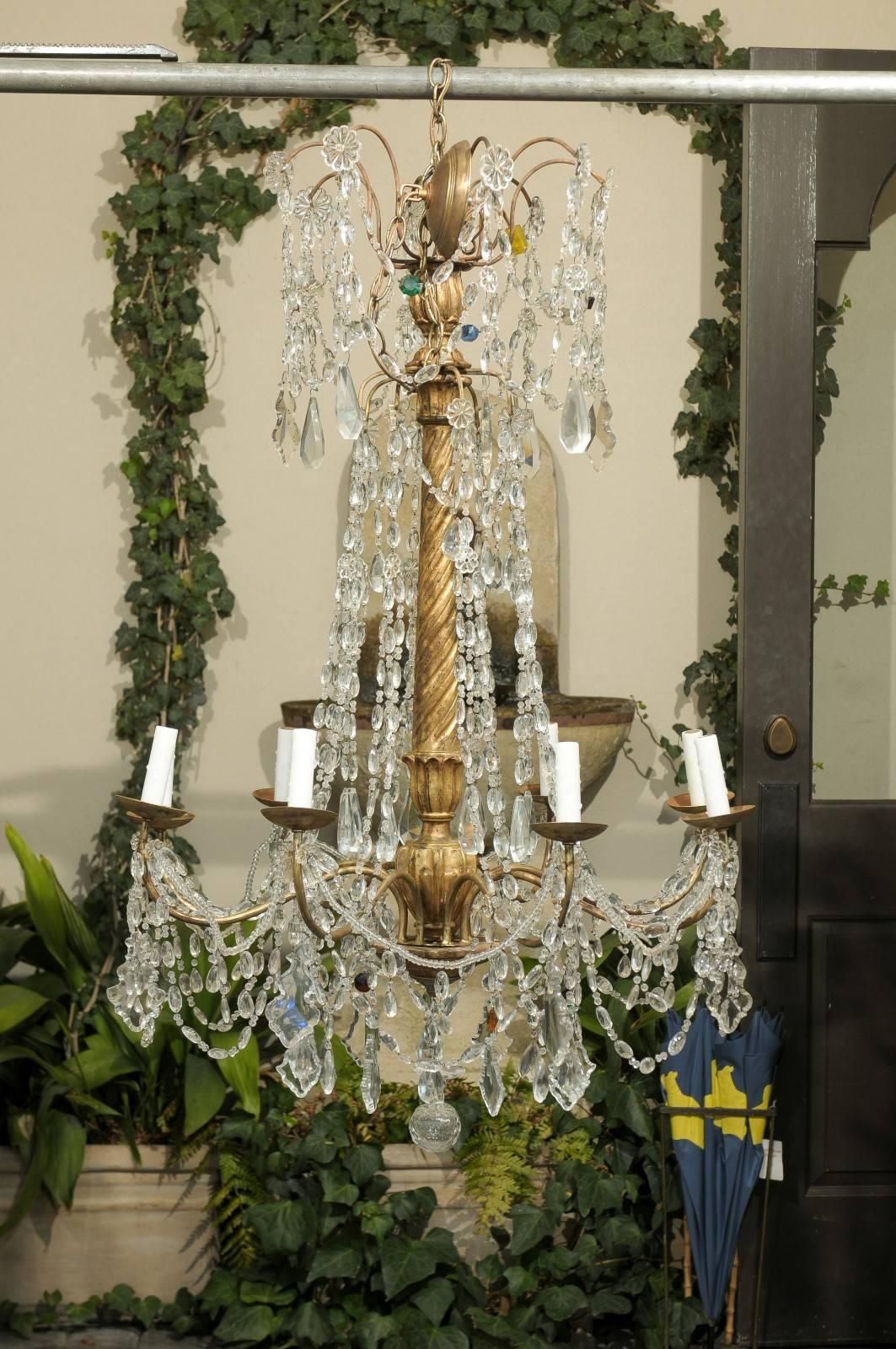 This Italian eight light crystal chandelier features a gilded and twisted wood central column with floral motif. The upper section is adorned with a variety of faceted crystals and rosettes. In the midst of these delicate ornaments, sits a green,