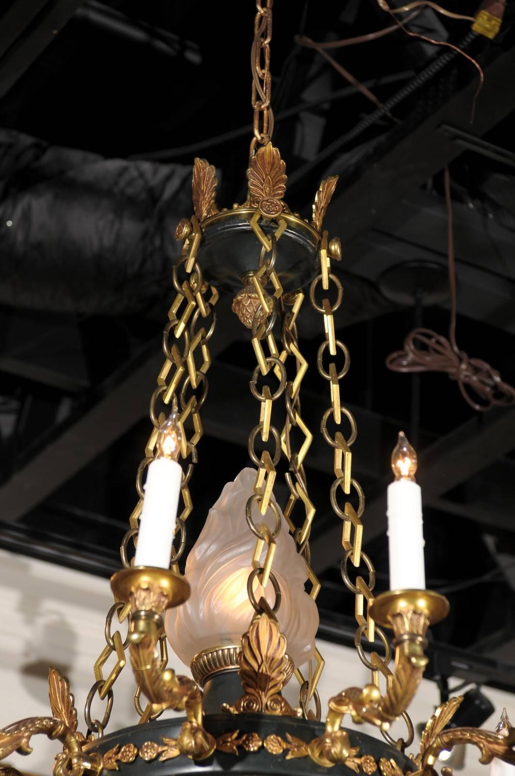 A French early 20th century Empire style ten-light chandelier with metal and brass accents. This French 1920s chandelier features a palmette adorned canopy in its upper section, connected to the central ring with profiled links. Ten scrolling arms,