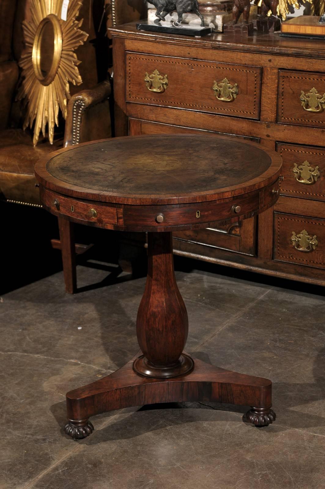 A regency leather top drum table from early 19th century England. This early 19th century Regency drum table features a circular top with leather inset over three drawers (and three faux drawers) with two brass knobs flanking a brass insert keyhole