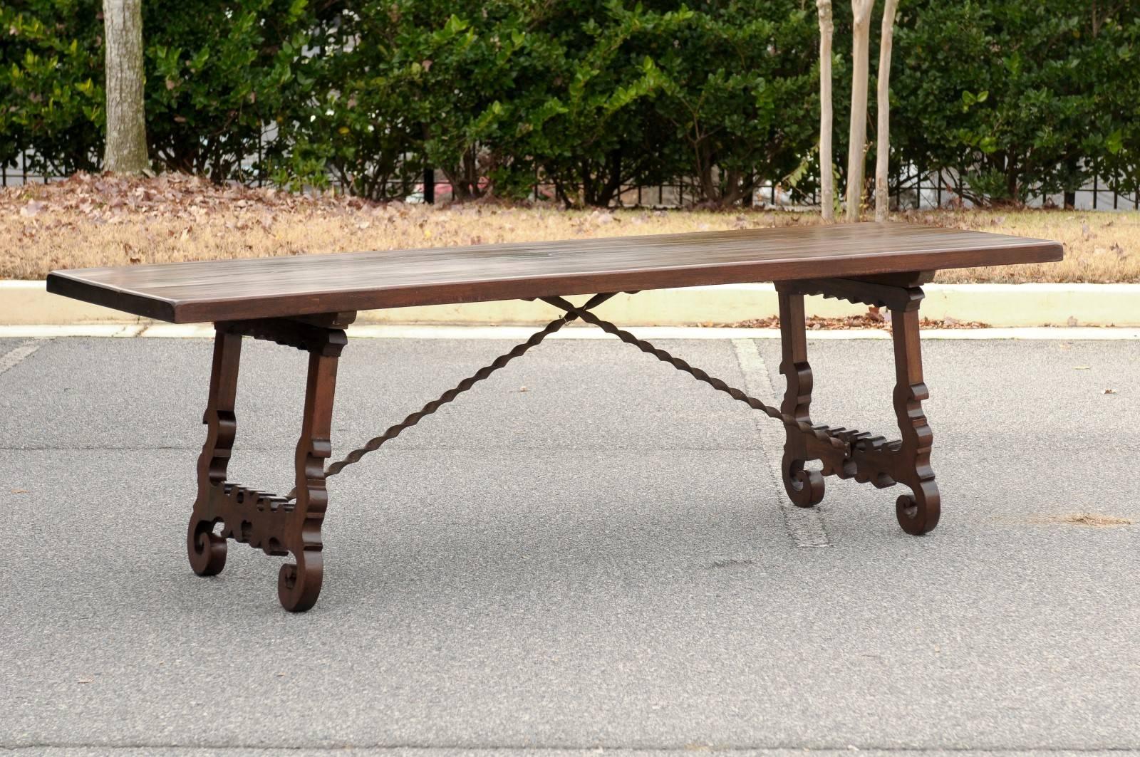 This is a late 19th century Long Italian trestle table with two Baroque style flattened lyre-shaped legs and a thick solid wooden single plank top, possibly made of walnut. The splayed double scroll carved legs are joined together by beautifully