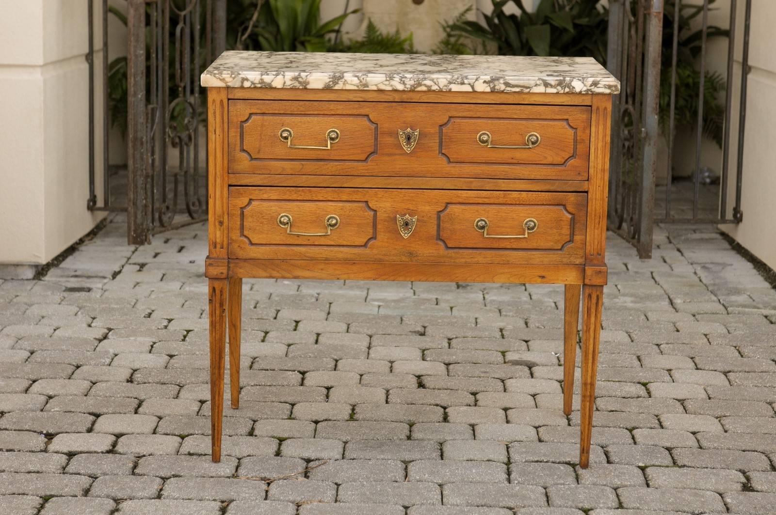 A French Neoclassical style oak two-drawer commode with marble top from the late 19th century. This French commode features an elegant variegated marble top, sitting above two dovetailed drawers. Each drawer is adorned with a brass shield-shaped