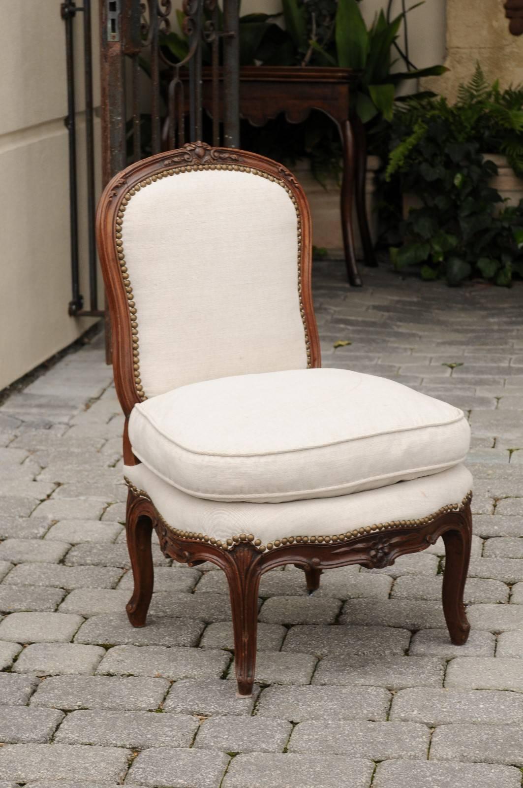 This French 18th century Louis XV slipper chair features a slightly slanted back with a delicately carved floral carving at the crest. This small size chair is upholstered with linen and nailhead surround on the back and seat with a comfortable