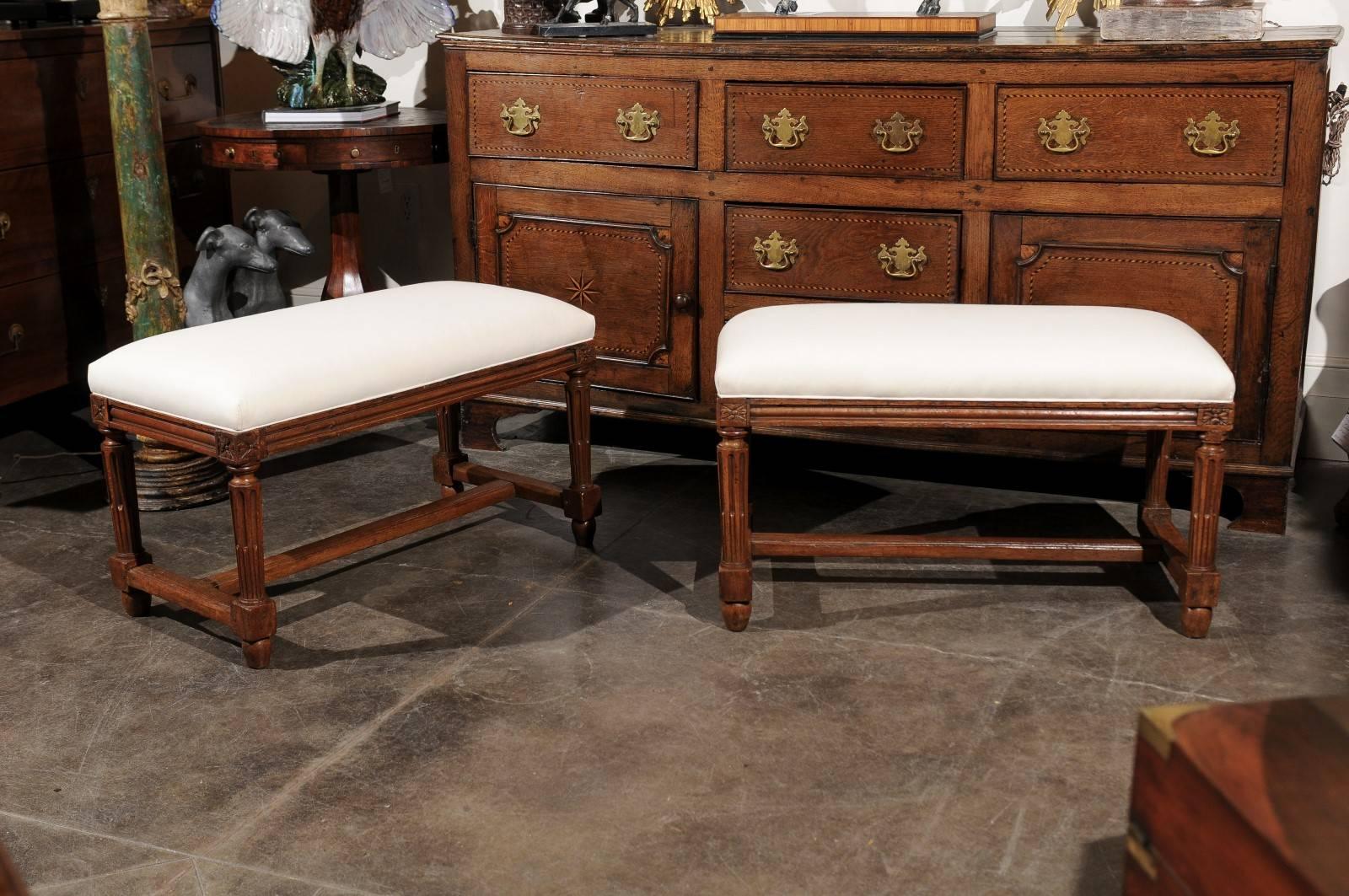 This pair of Italian small size wooden benches features an upholstered seat over a nicely carved walnut base. The reeded skirt is adorned with a rosette placed in each corner. The benches are raised on four fluted and reeded legs, joined by a cross