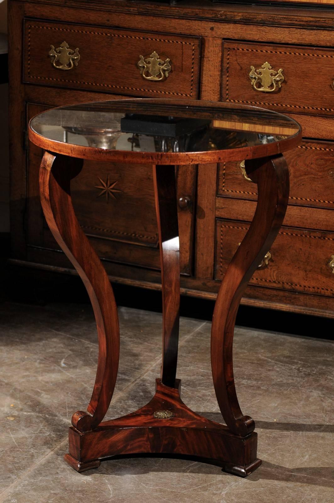 This French 19th century Restauration style guéridon side table features a circular distressed mirrored top supported by three s-scroll shaped legs. These elegant legs are raised on a triangular base, showing a concave profile on all sides. The