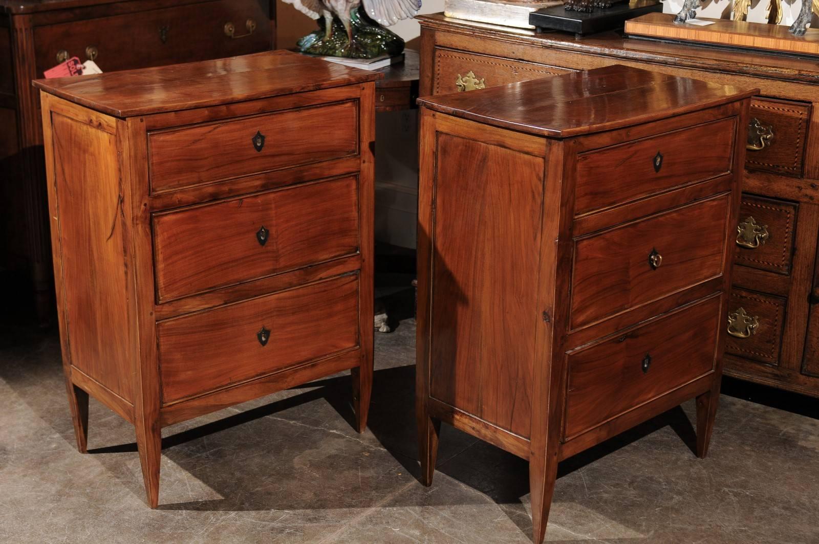 This pair of French Directoire style small commodes from the early 20th century are made of cherrywood. A rectangular top sits over three drawers adorned with a central escutcheon shaped as a shield. The sides are made of recessed panels. The