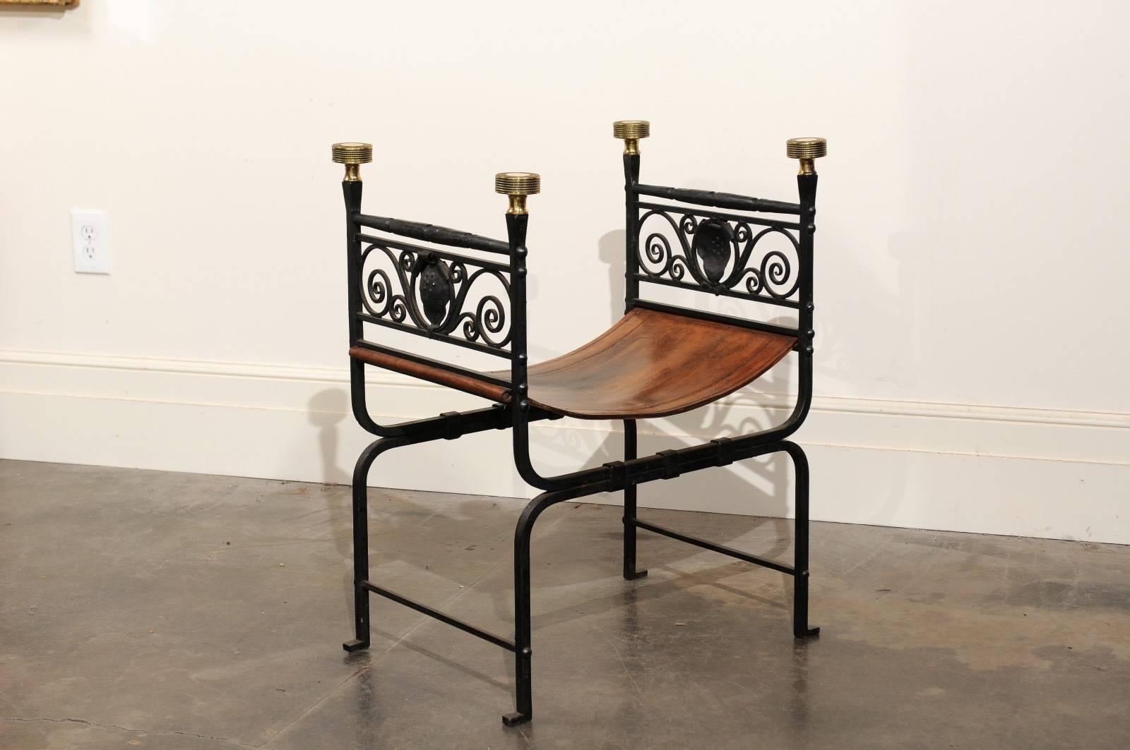 This stylish English Campaign stool from the turn of the century features an H shaped frame securing a brown leather seat. The eye is immediately drawn to the exquisite black iron frame, adorned on the sides with a delicate décor of volutes flanking