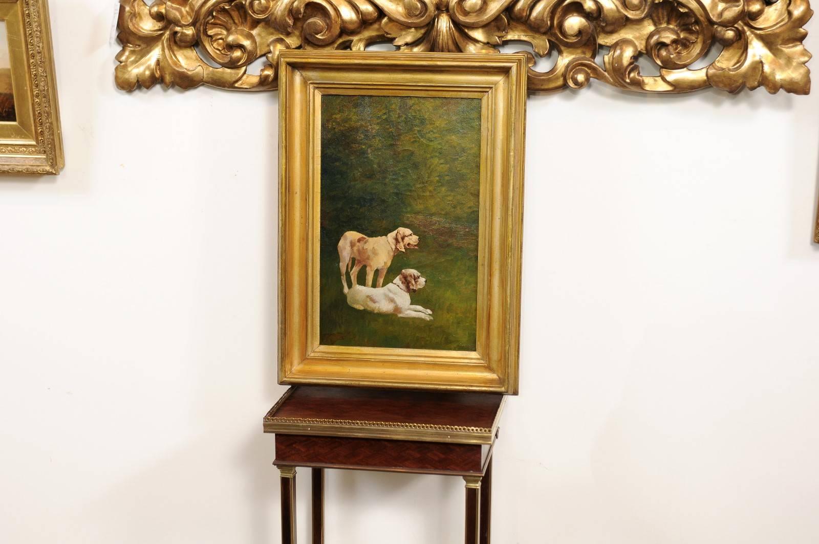 This French vertical format oil on canvas dog painting from the early part of the 20th century features a simple composition: two dogs with light coats in the center. One is standing while the other one is peacefully resting on the grass. The