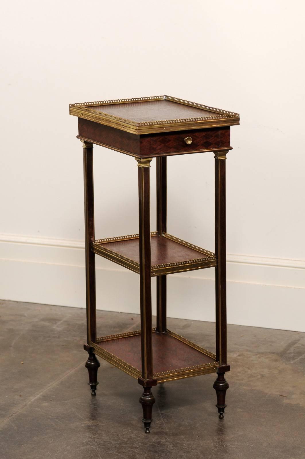 This English mahogany étagère from the late 19th century features three parquetry decorated shelves surrounded by a delicate brass gallery. The upper shelf contains a single drawer adorned with parquetry inlay. The ensemble is secured by thin square