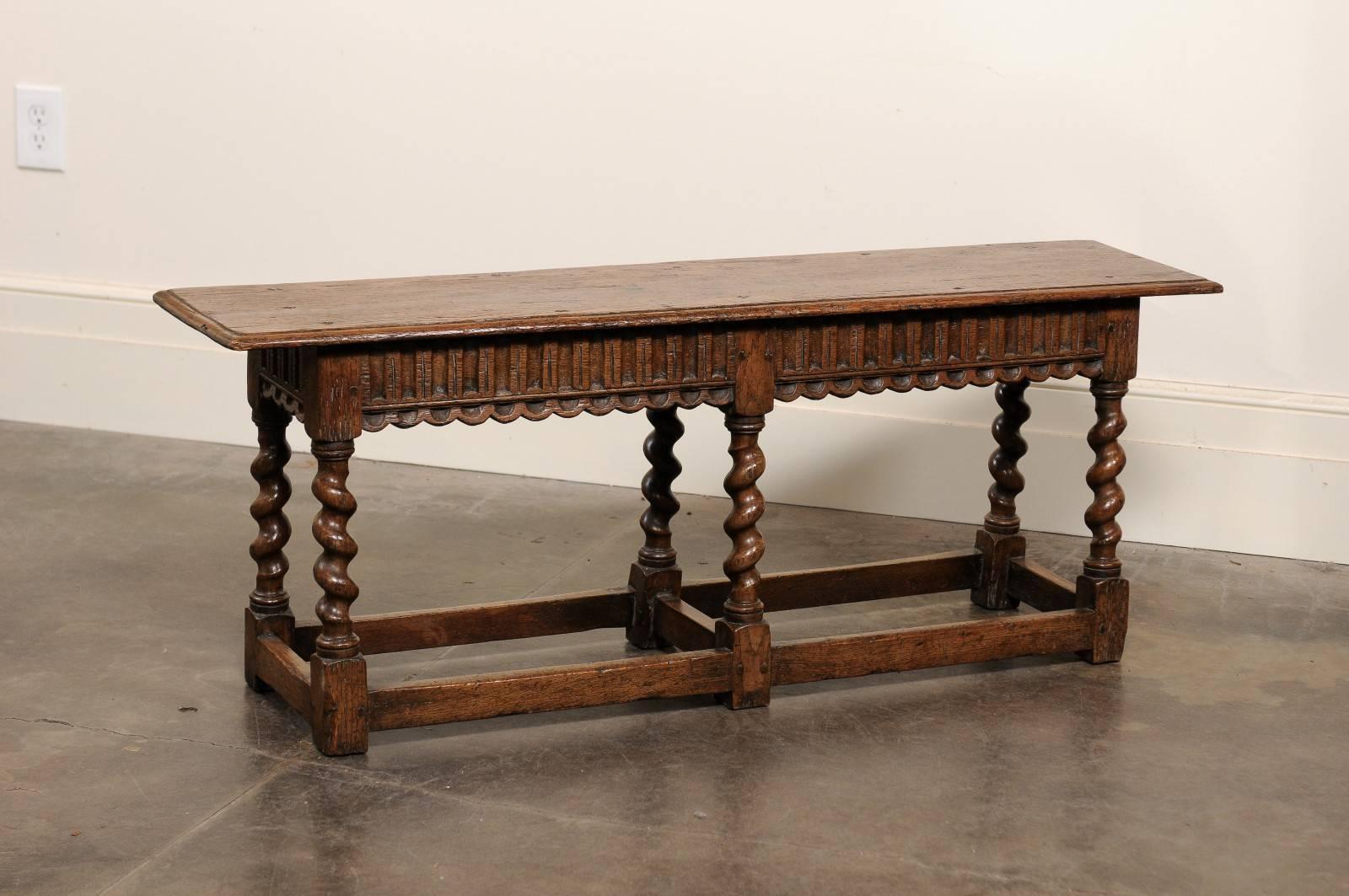 This English carved oak backless bench from the early 20th century features a rectangular wooden seat with beveled edges over a nicely carved seat rail. Six stylish barley twist legs support the ensemble and are connected to one another by side