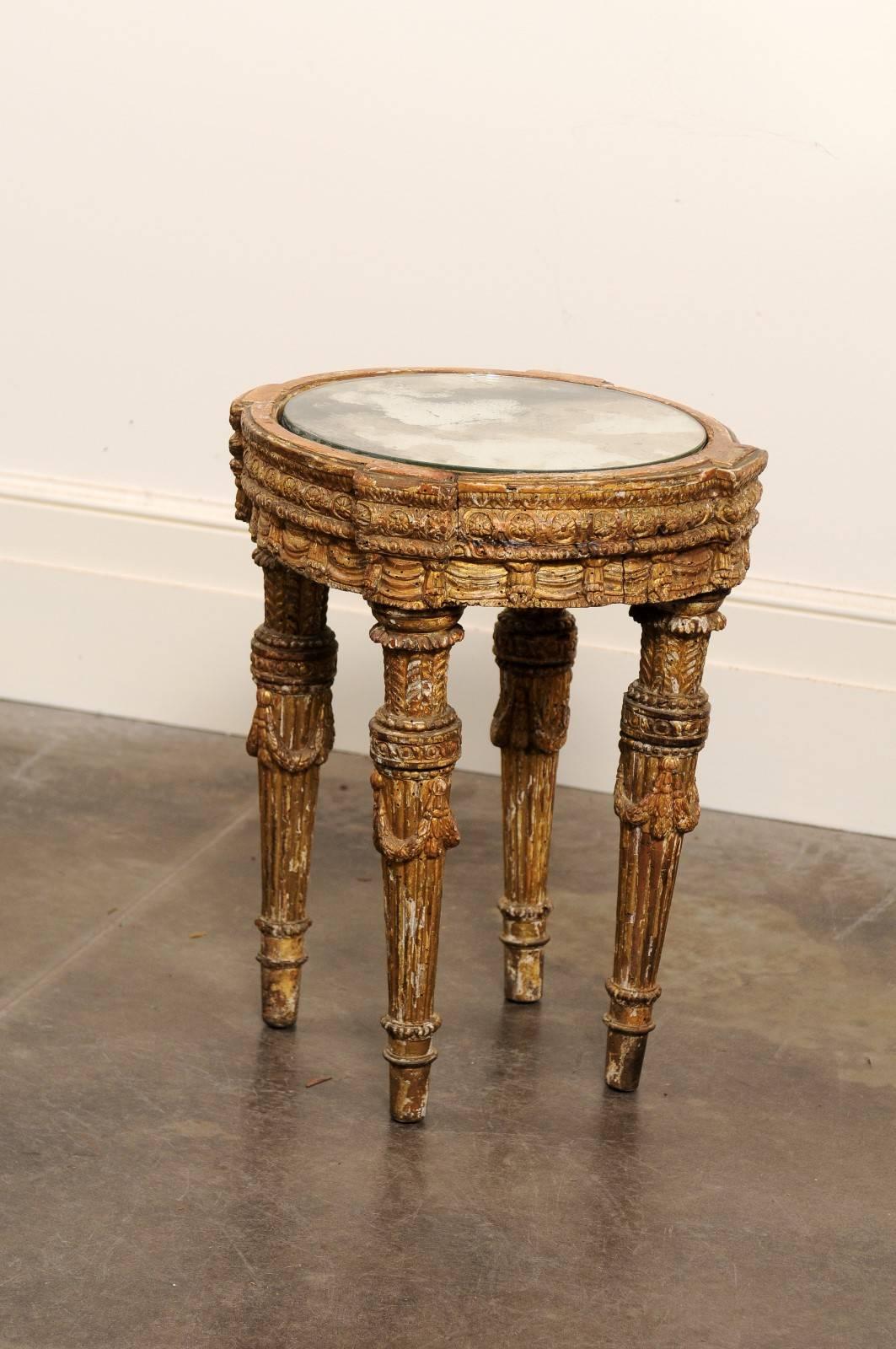 This Italian mid-18th century petite drinks table features a circular top with distressed mirror over a richly carved wooden base. The apron is adorned with various motifs such as egg-and-dart and rosettes in medallions. Four carved fluted tapering