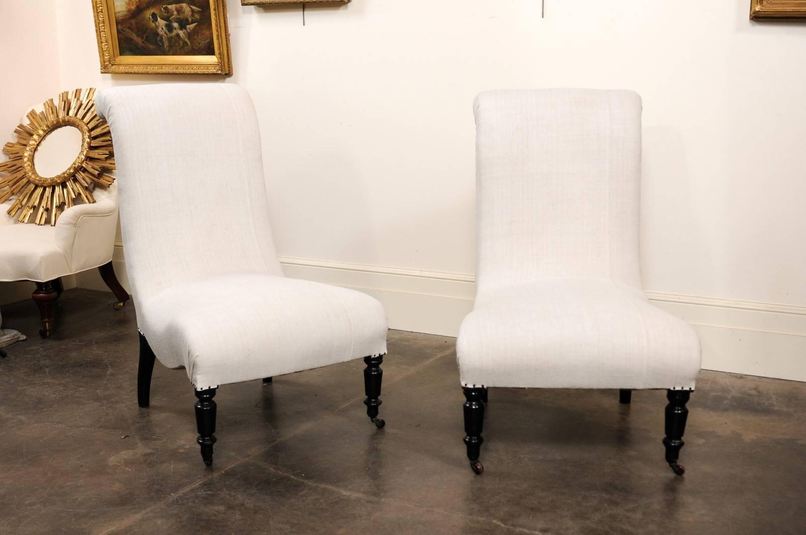 This fabulous pair of English upholstered slipper chairs from the turn of the century (19th-20th) features out-scrolled backs over nicely curved seats. The chairs are raised on four ebonized oak feet, two turned with casters in the front and two