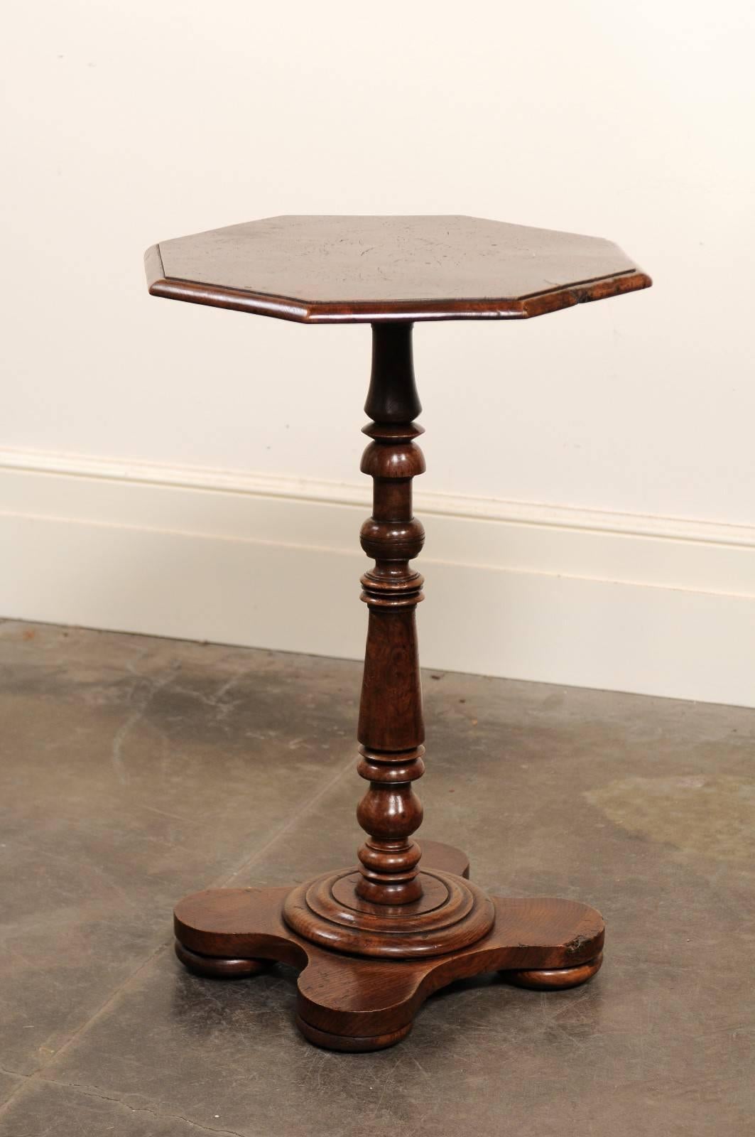 This petite English burled elm side table from the late 19th century features an octagonal top over a turned central pedestal. The base, reminiscent of a quatrefoil motif, is raised on four bun feet. The richness of the burled wood on the top,