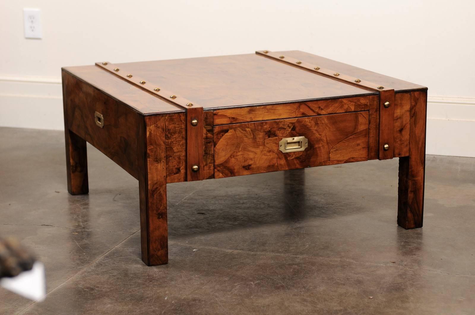 This English Campaign coffee table from the mid-20th century features an exquisite burled wood top over a single drawer. The top is adorned with two wooden bands with brass nailheads, reminiscent of the leather straps of a trunk or suitcase. The