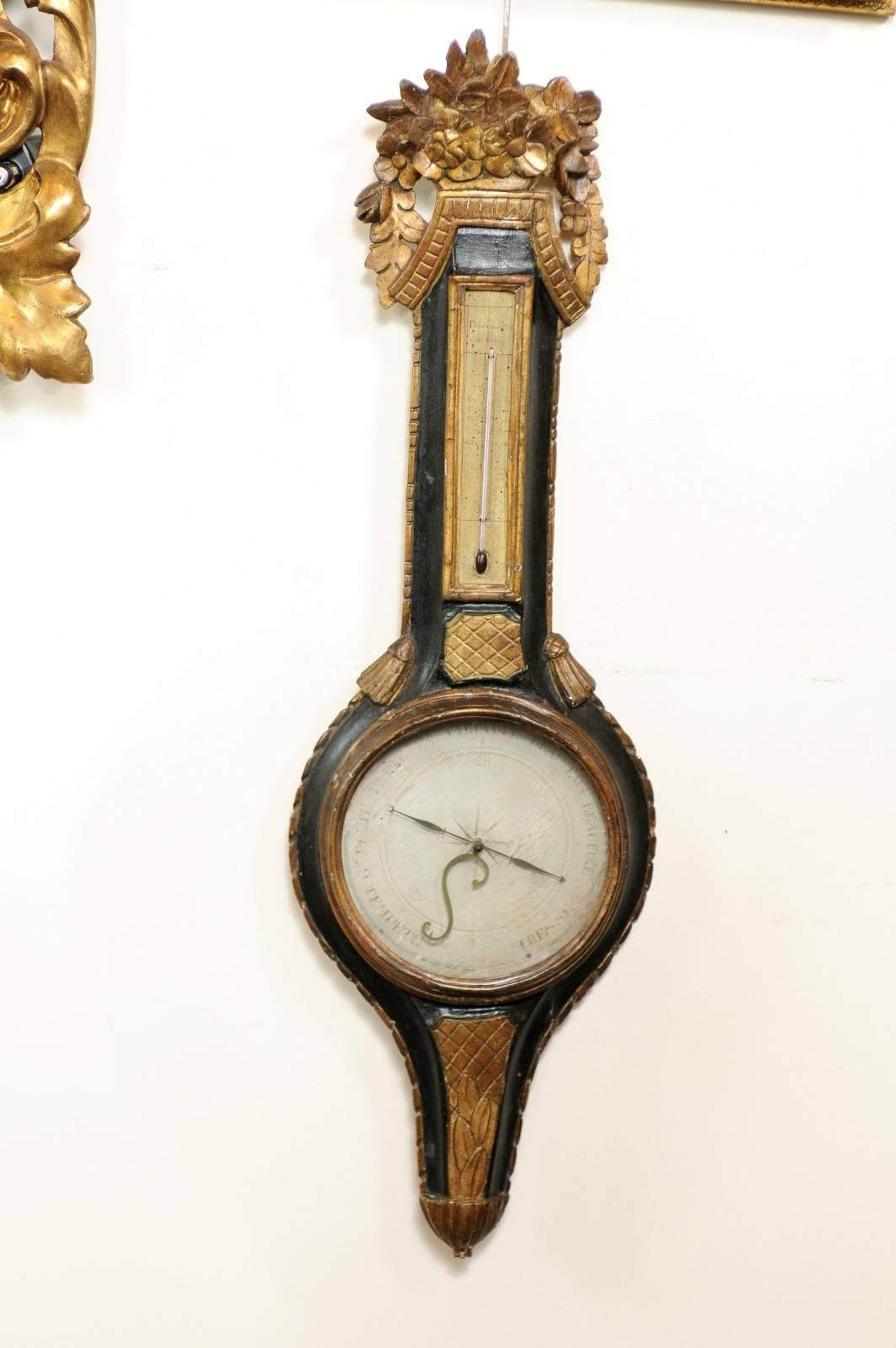 Dieses Barometer und Thermometer im französischen Louis XVI-Stil besitzt eine exquisite geschnitzte Goldholzkrone mit zarten Blattwerkmotiven. Unter dem Wappen befindet sich das Thermometer, das von einem schwarz lackierten Gehäuse umgeben ist, auf