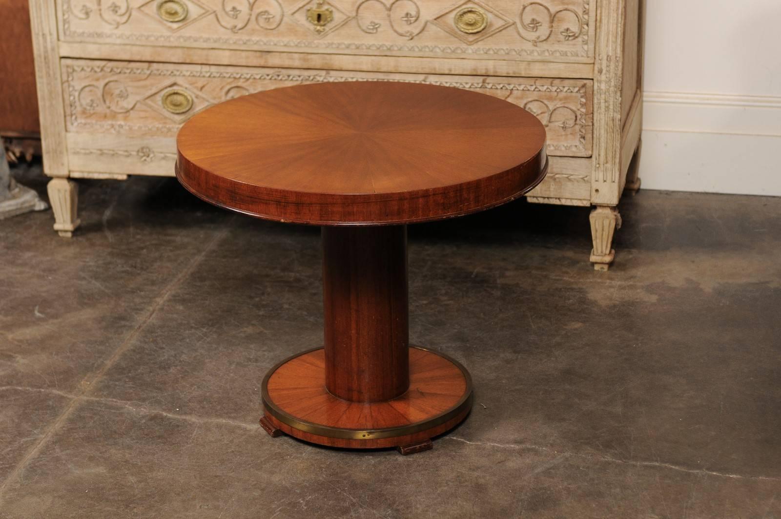 This petite French mahogany veneered low side table from the mid-20th century features a circular top over a pedestal base. This base is raised on a round pediment of smaller size, surrounded by a brass ring and resting on four feet. The top is
