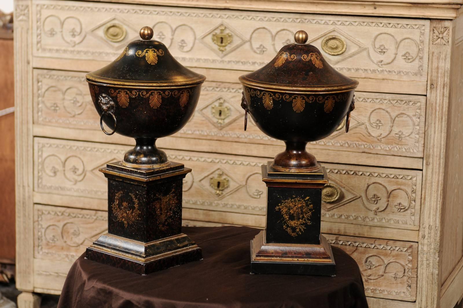 This pair of French tole urns from the early 20th century is made of black circular vessels decorated with gilded palmettes and topped with black and brown mottled lids. Each urn is adorned with ring pulls on the side secured on gilt metal lion back
