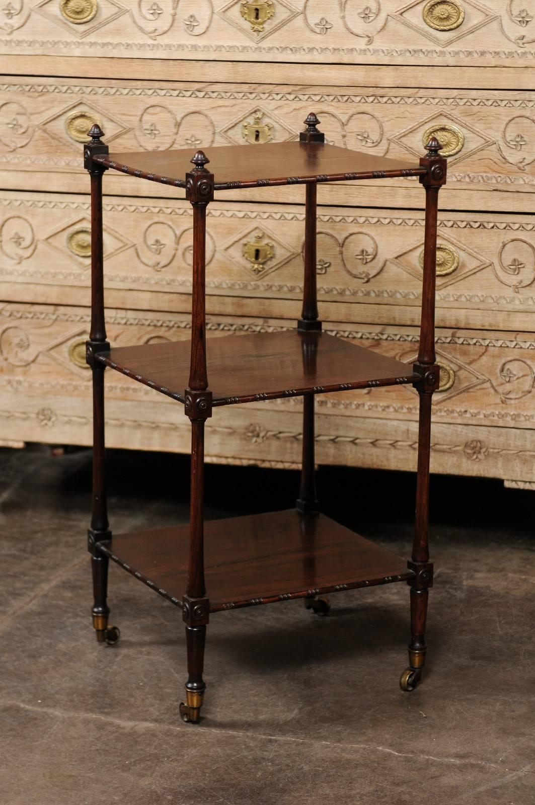 This English rosewood tiered trolley from the mid-19th century features three rectangular shelves with carved edges on four sides secured within four elegant side posts. The posts are topped with finials and adorned with block joints with