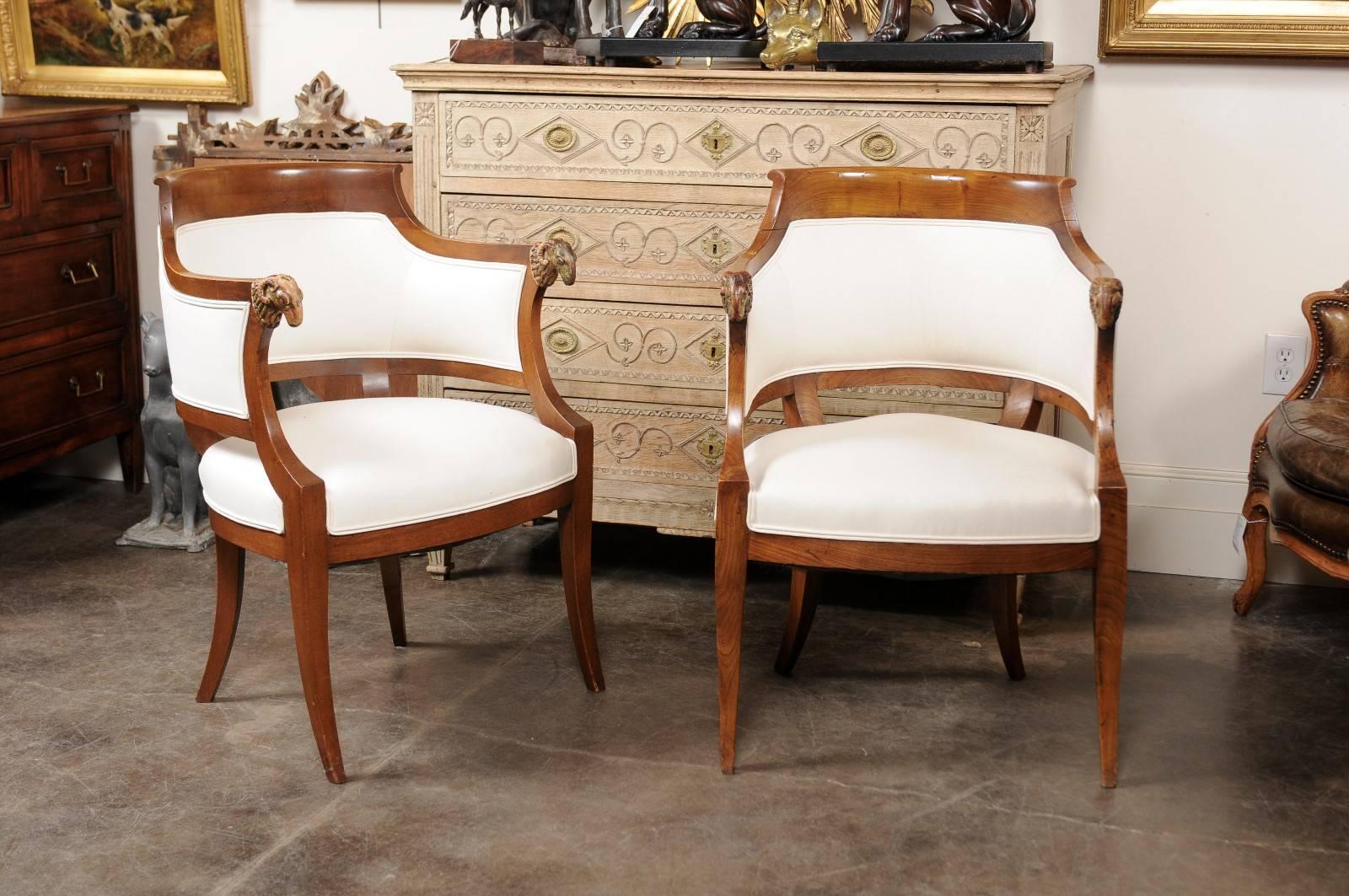 This pair of Austrian Biedermeier armchairs from the mid-19th century features upholstered barrel backs adorned with exquisite carved ram's heads on the arms. Skillfully depicted with great details, these are the only decorative accents that the