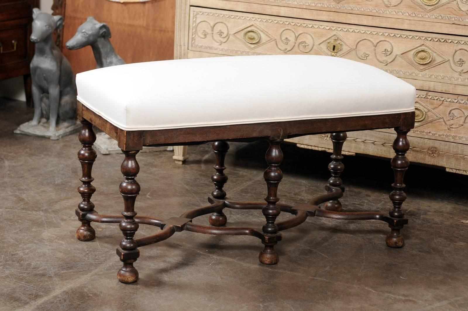 This French Louis XIII style oak backless bench from the late 19th century features a rectangular smooth muslin upholstered seat over six exquisite turned legs. These legs are connected to one another through curved X-shaped stretchers with diamond