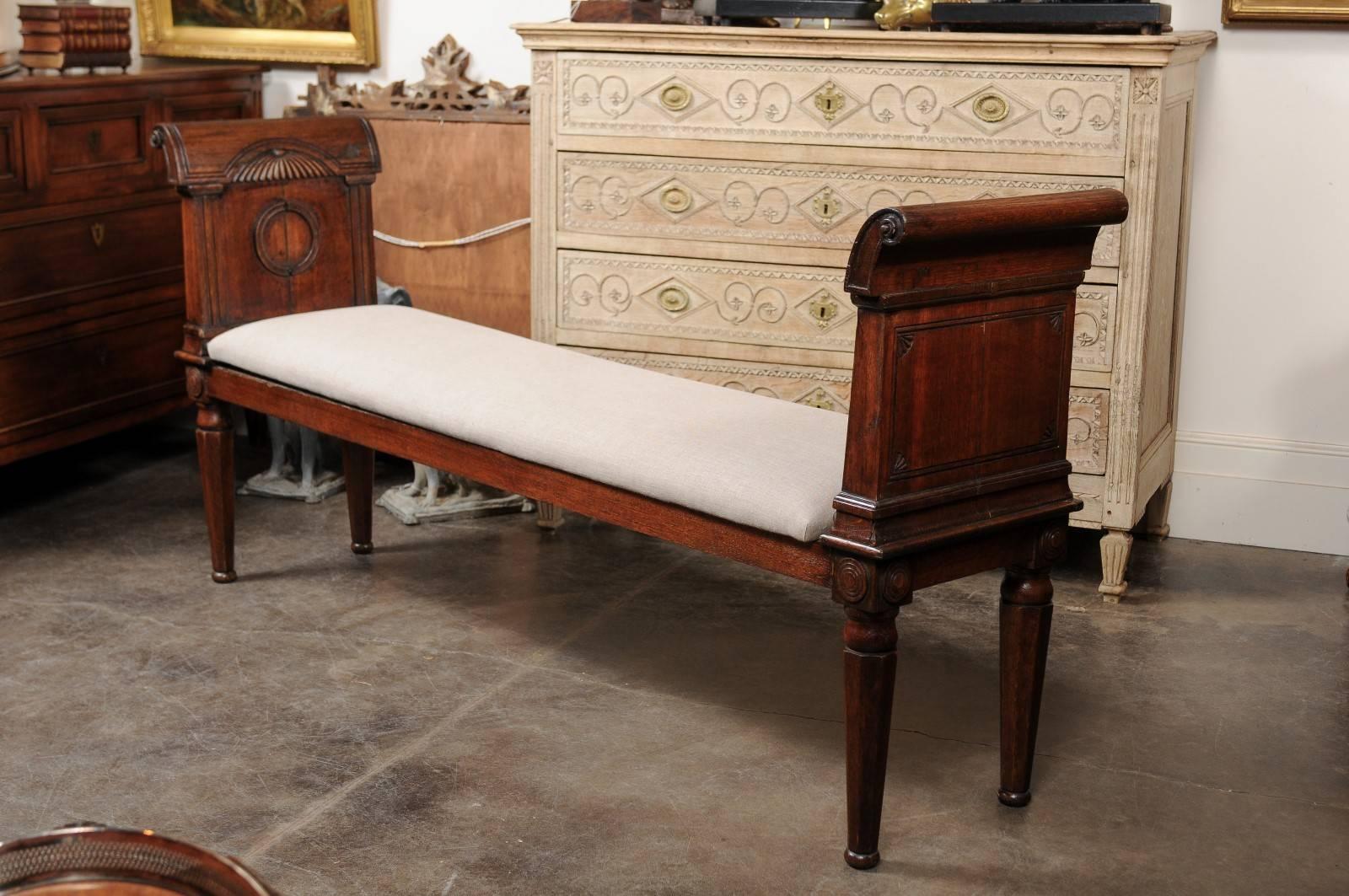 This English oak backless bench from the late 19th century features two beautiful out-scrolled arms with carved motifs flanking a long rectangular upholstered seat. Paying great attention to details, the maker decorated the arms' back panels with