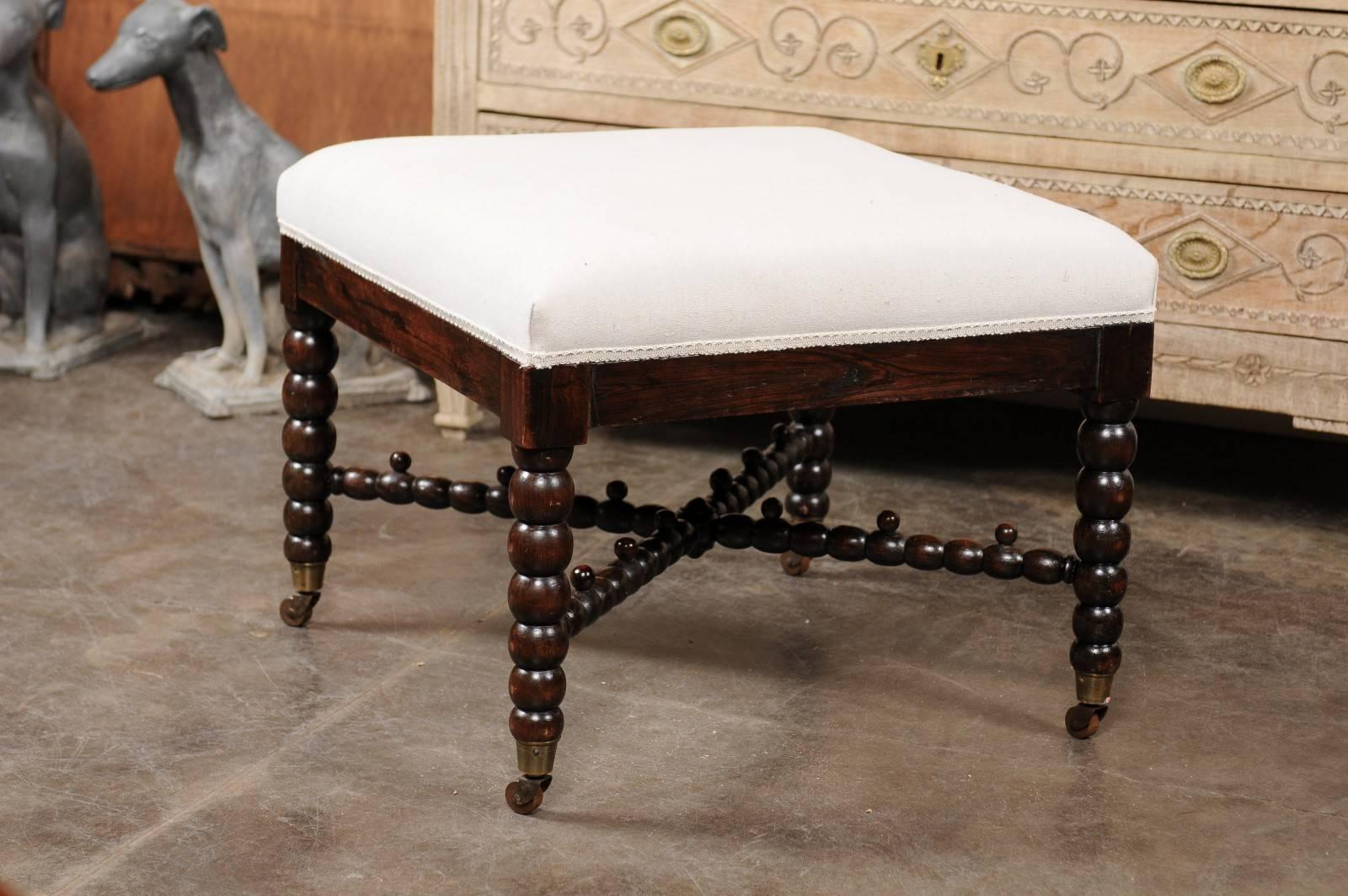 An English bobbin leg ottoman with upholstered seat. This English wooden ottoman from the late 19th century features a square seat covered with a smooth muslin upholstery over four exquisite bobbin legs raised on casters. An unusual cross stretcher