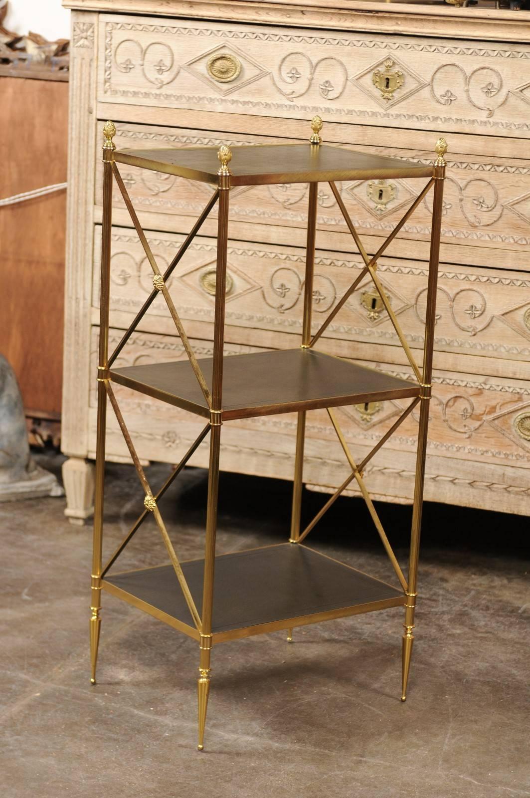 This Italian Maison Jansen style Mid-Century Modern tiered stand features a brass frame delicately flanking three black leather shelves. The upper shelf is adorned in each corner with four brass pinecone finials while the lower shelves are secured