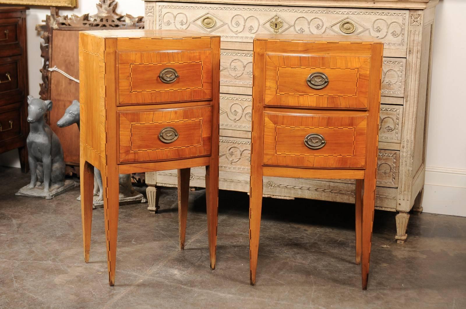 This pair of Austrian Biedermeier petite commodes from the mid-19th century features exquisite veneered tops with rectangular banding over two small drawers. Each central oval mounted plate with bail handle is surrounded by a delicate banding,