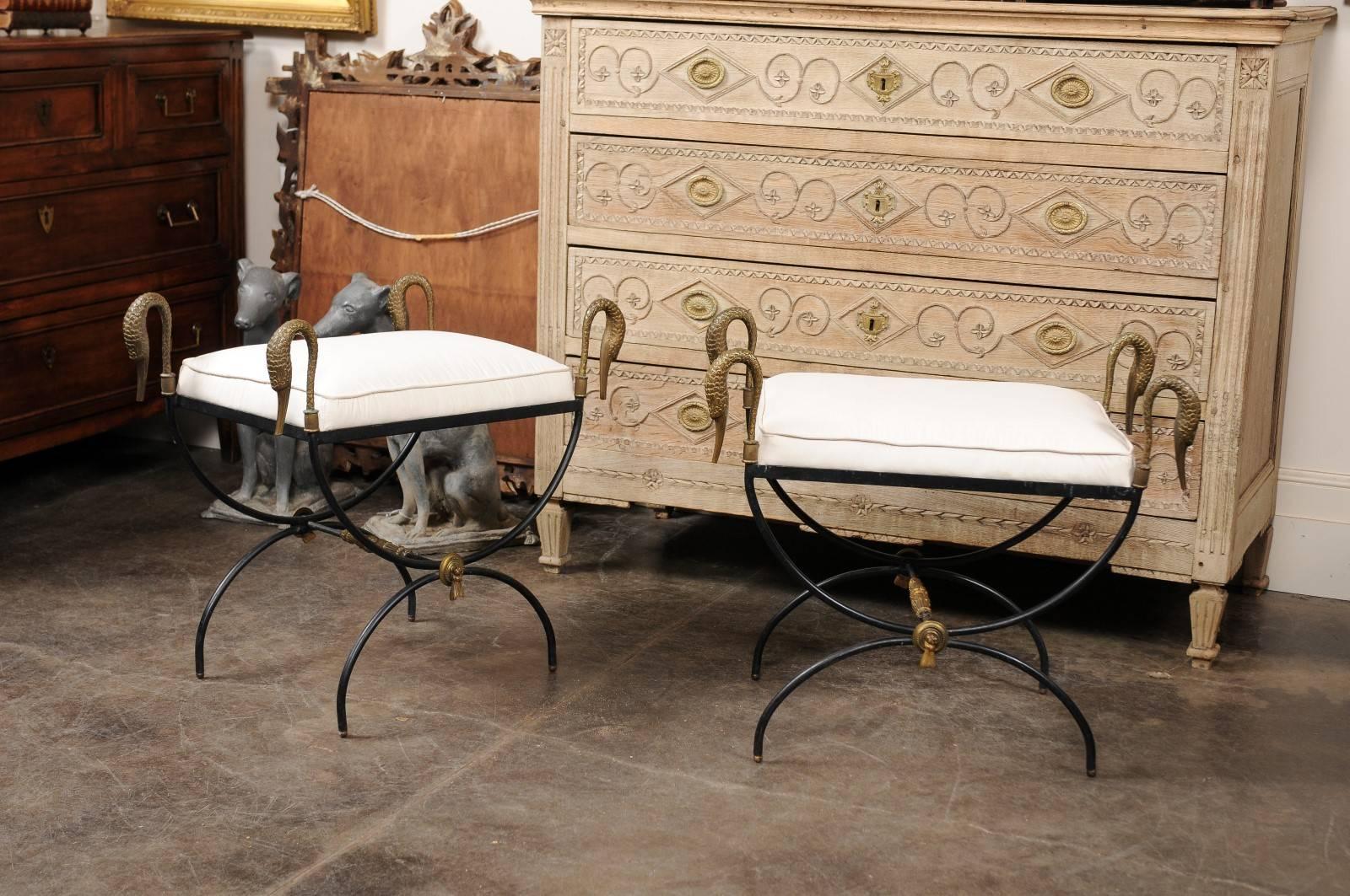 This pair of French neoclassical style stools with brass swans from the mid-20th century features rectangular seats upholstered in a smooth muslin, supported by wrought iron Curule style bases with cross stretchers. The arms are adorned with