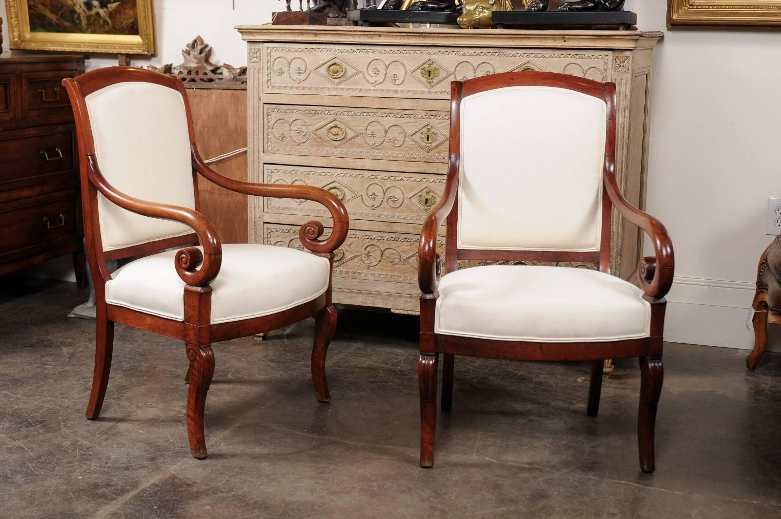 This pair of French Empire style walnut fauteuils from the mid-19th century features slightly out-scrolled backs with beautifully scrolled arms. The armchairs are raised on four legs each, two cabrioles - reminiscent of the Louis XV era - in front,