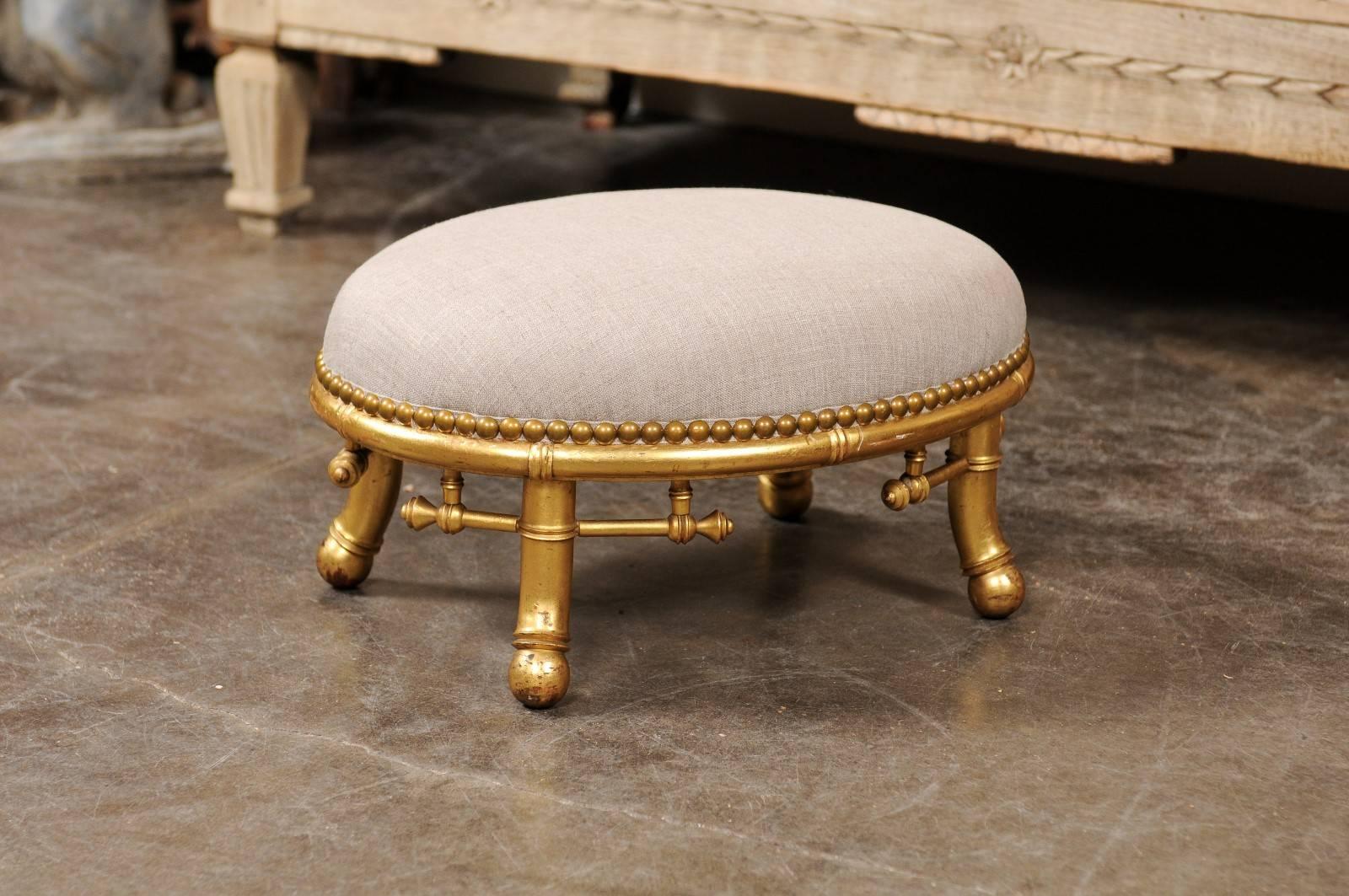 This English faux-bamboo petite footstool from the turn of the century (19th-20th century) features an oval muslin upholstered oval seat with nailhead trim, over a giltwood frame. Raised on four short splayed feet, this adorable English footstool,