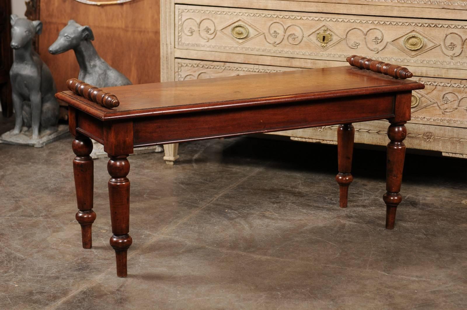 This English mahogany hall bench from the late 19th century features a rectangular wooden seat with beveled edges adorned with two exquisite turned arm supports. The piece is raised on four elegant turned legs with tall cylindrical feet. This bench