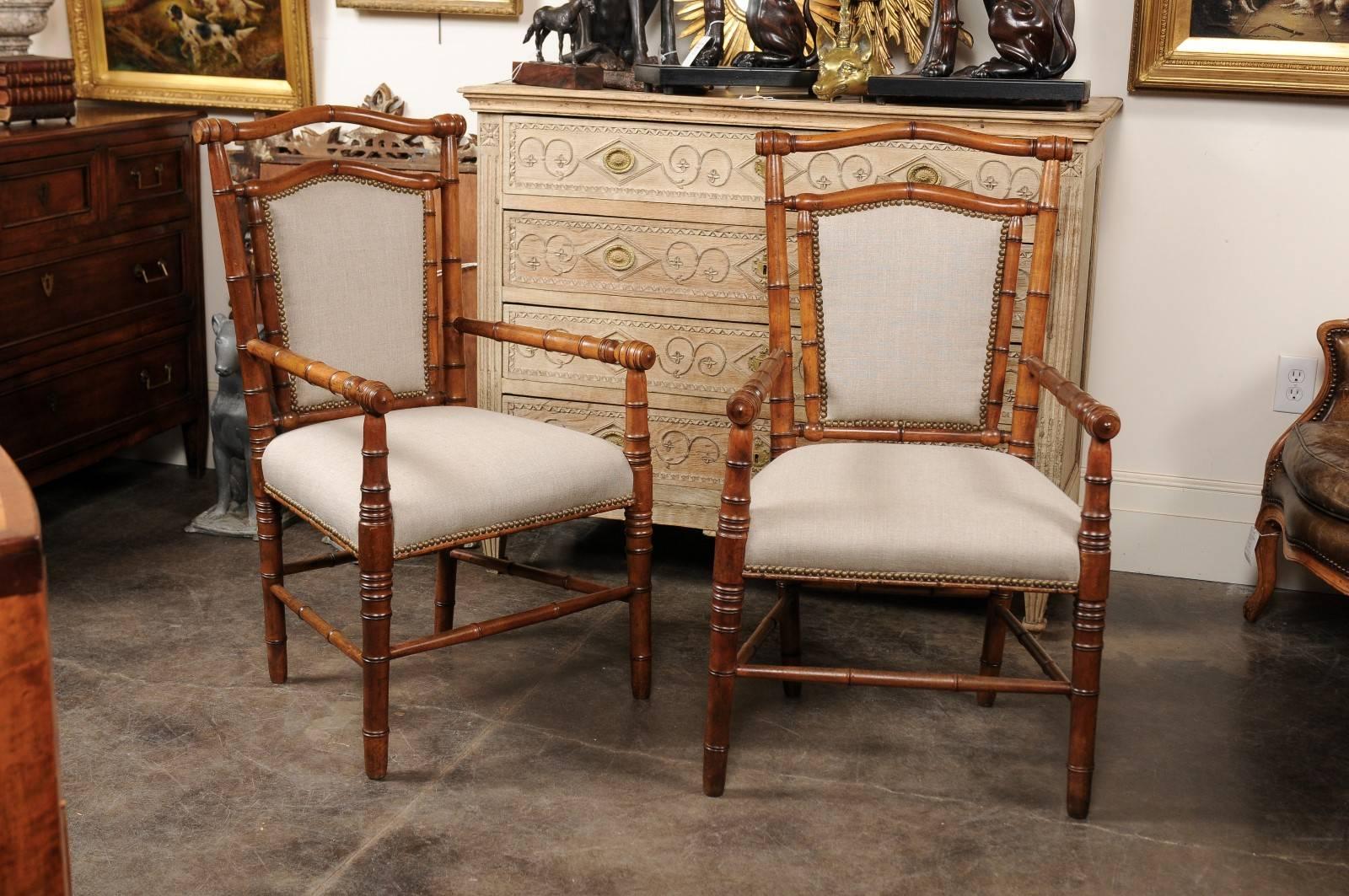 This pair of English faux-bamboo chairs from the turn of the century features slightly slanted backs with partial upholstery and nailhead trim over turned legs with side stretchers. The straight lines seen on the arms, legs and stretchers are gently