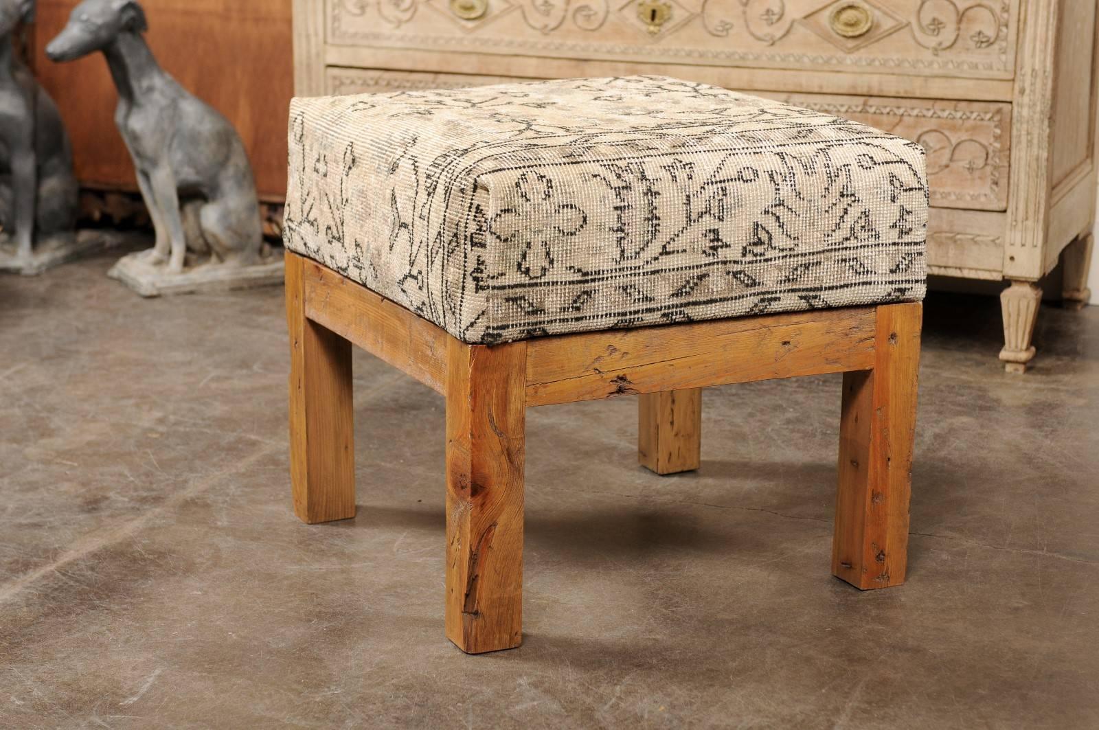 This wooden stool features an upholstered seat covered with a vintage Turkish rug over an old wooden base also from Turkey. The comfortable seat features a light color tone adorned with various geometrical patterns, and is raised on four straight