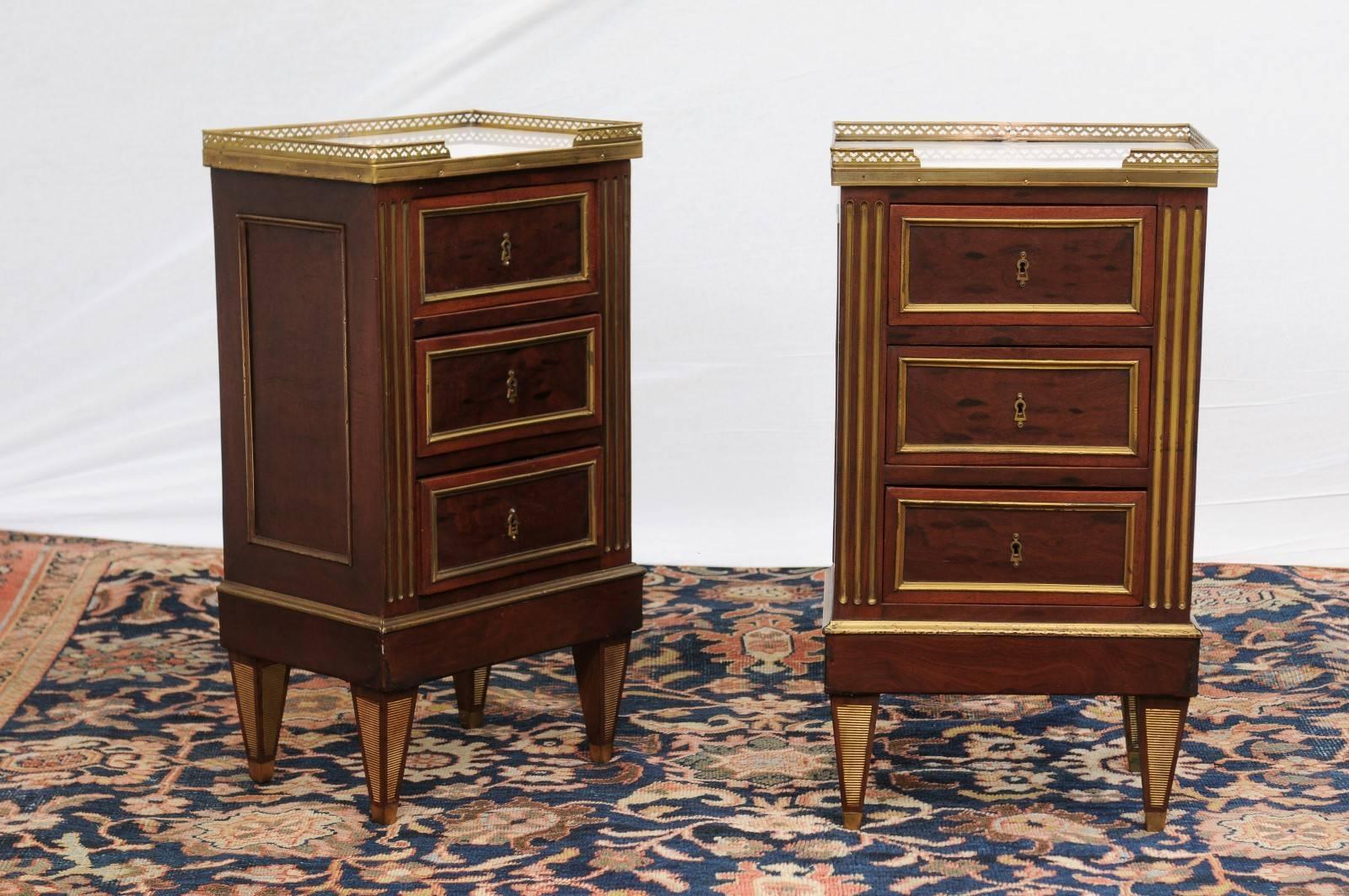 This pair of French Directoire style petite commodes was made in the early 20th century. Each features a three-quarter brass gallery with heart-shaped motifs surrounding a white marble top. The body is made of three dovetailed drawers with gilded