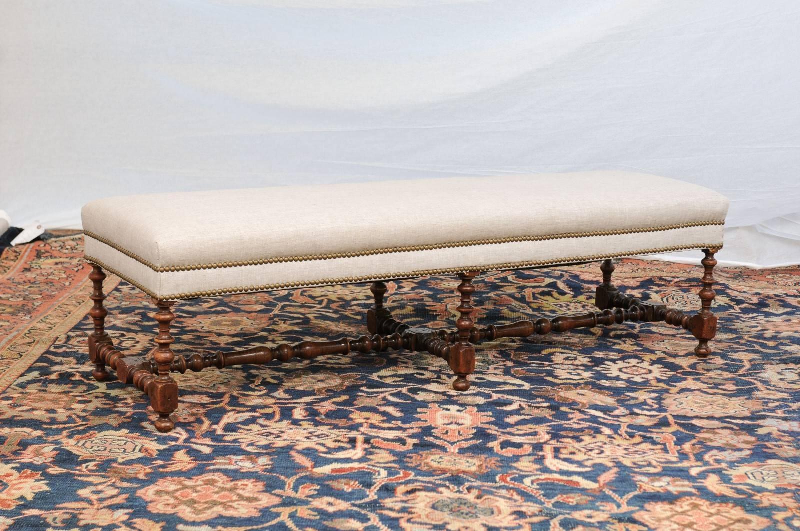 This English upholstered backless walnut bench from the early 20th century features a long upholstered seat with double nail head trim on the surround. The top is raised on six beautiful bobbin legs connected to one another with a cross stretcher.