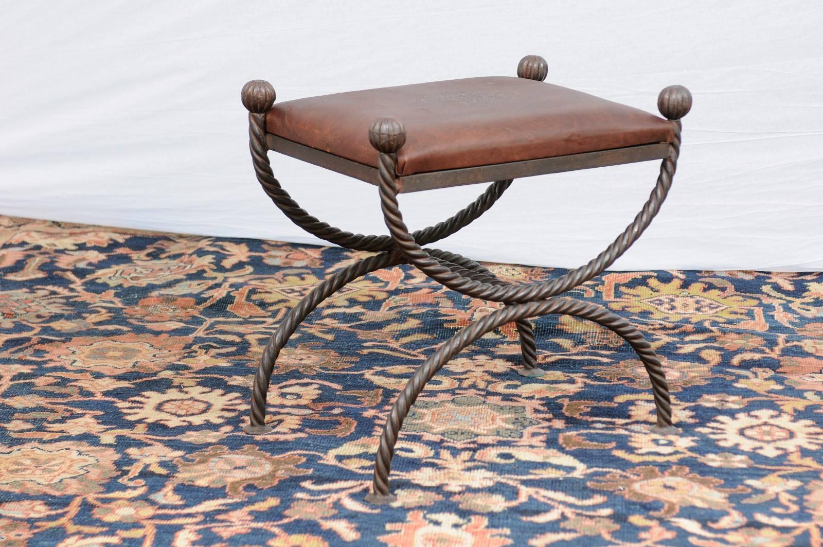 This English Curule style stool from the mid-20th century features a rectangular brown embossed leather seat over a savonarola rope style iron base. Each corner of the seat is marked with a round finial, adorned with gadroon shaped motifs. The