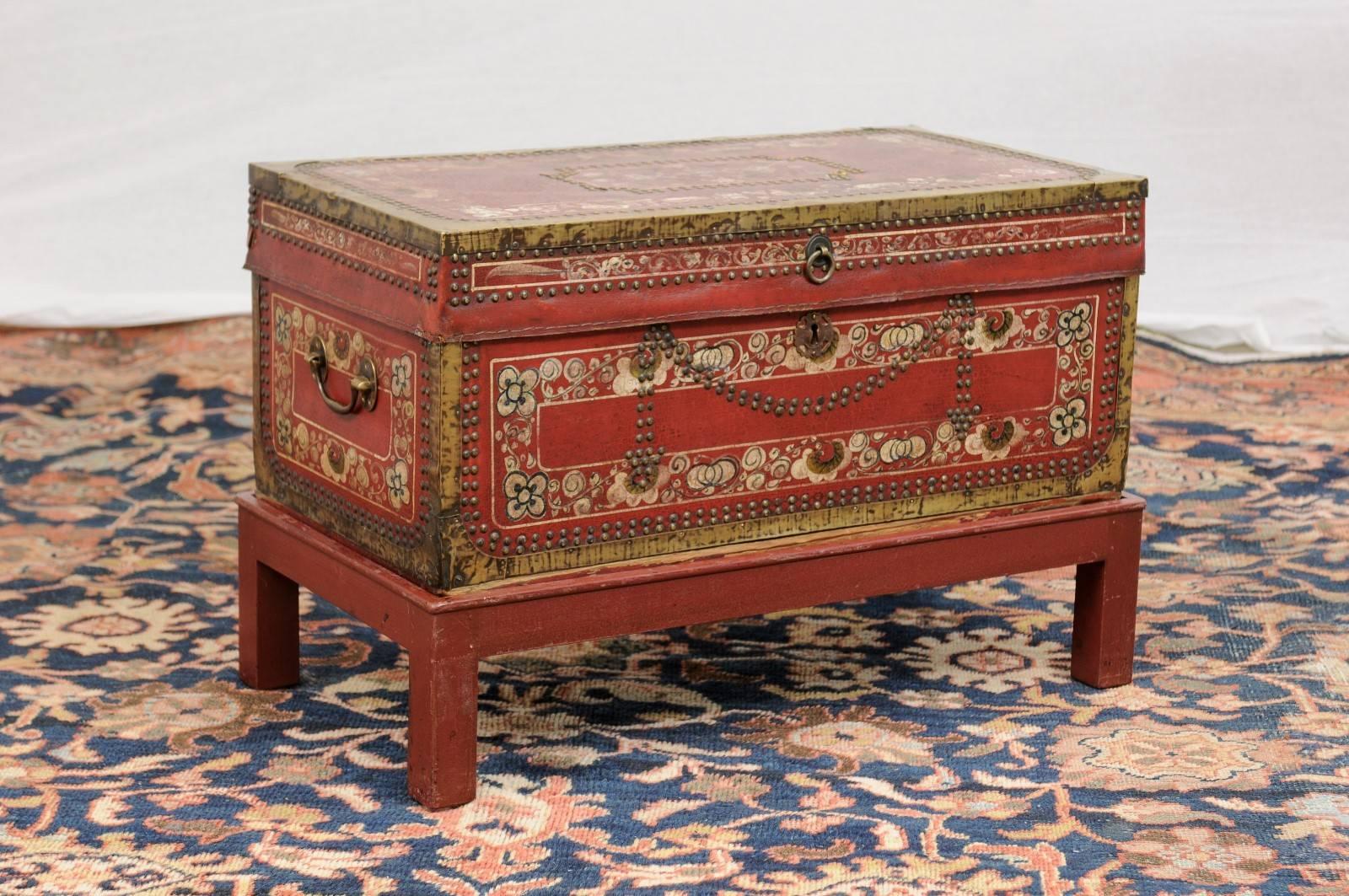 This English camphor wood trunk on stand from the late 19th century features a painted leather body with brass accents in the corners and nailheads. The red background is adorned with white floral motifs and darker accents. The nailheads on the