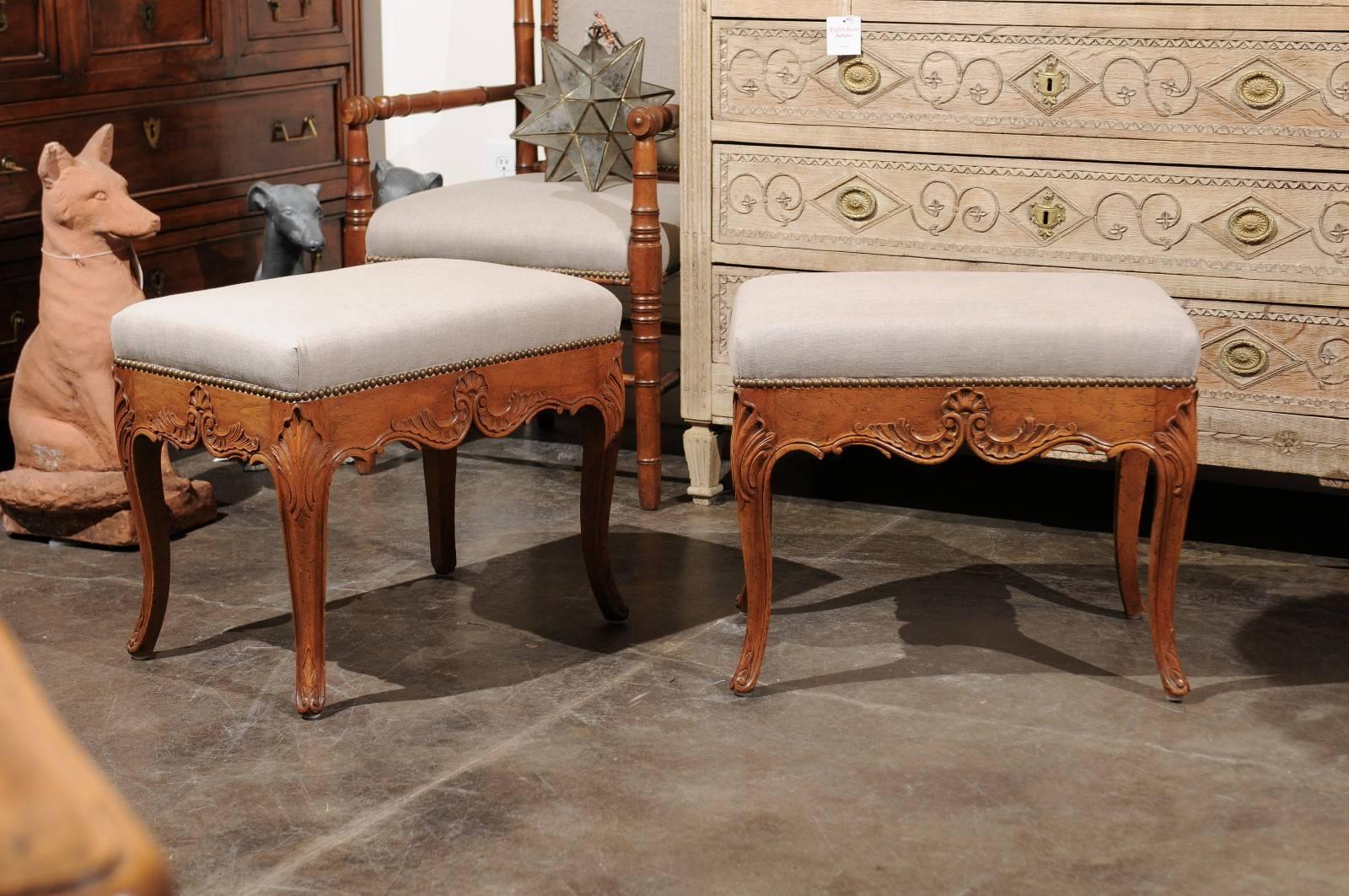 A pair of Italian Rococo style upholstered carved walnut stools from the mid-20th century. Each of this pair of Italian walnut stools features a rectangular seat upholstered in linen with nailhead trim, over a rocaille adorned scalloped seat rail.