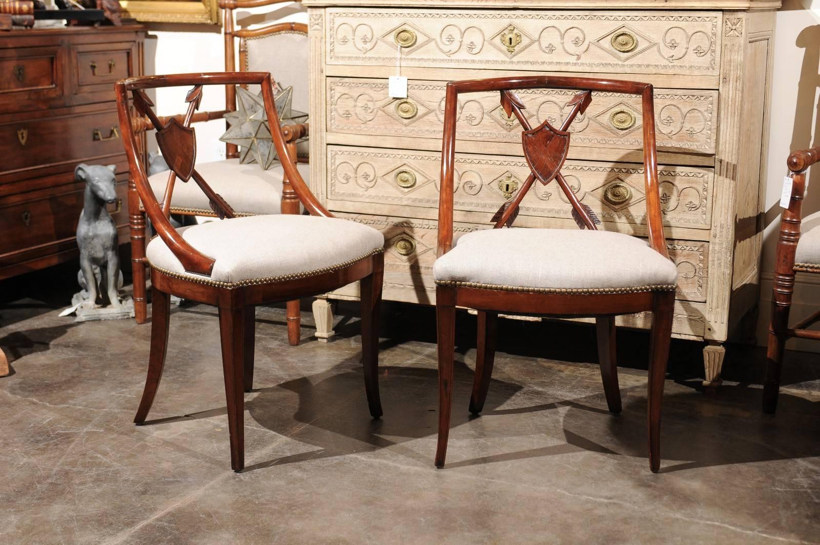 A pair of Biedermeier period Austrian upholstered armchairs with trophy of arms backs from the mid-19th century. Each of these Biedermeier armchairs circa 1840 features a sleek, curved back pierced in its center with trophy of arms made of a shield