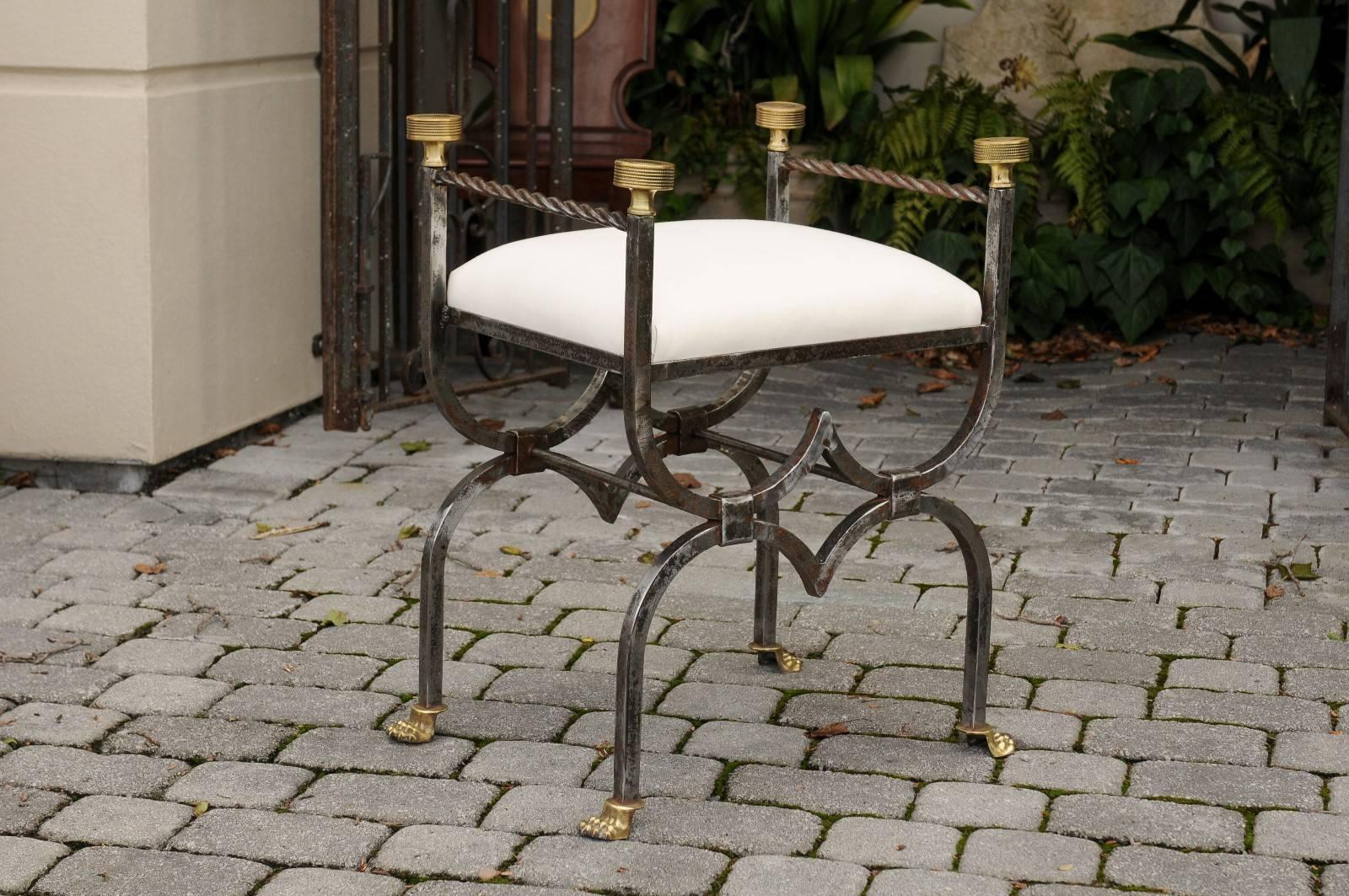 An English steel and brass upholstered stool from the turn of the century. This English stool circa 1900 features a modified curule-shaped body with double curvature, raised on brass lion paw feet and topped with beaded medallion finials of the same