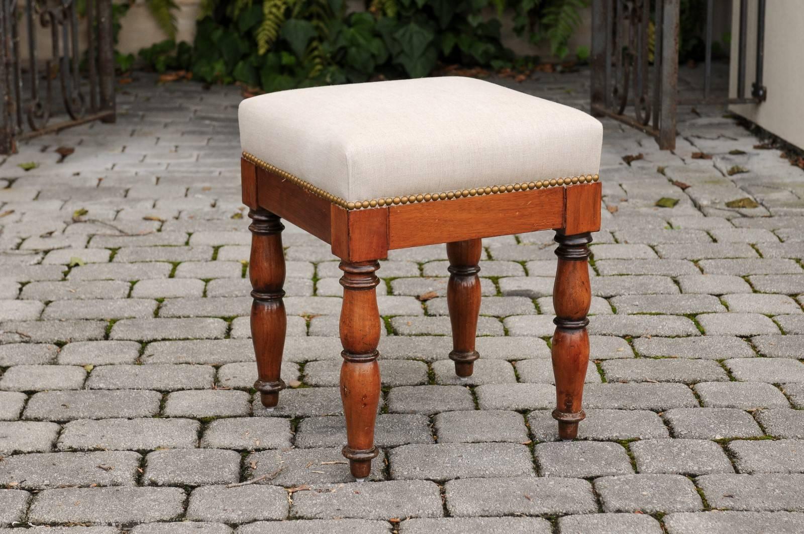 An English walnut stool with upholstered seat from the second half of the 19th century. This English wooden stool features a square seat covered in linen with brass nailhead trim. The simple skirt is raised on four turned legs, gracefully swollen in