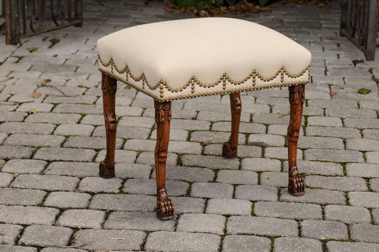 An English carved oak stool reupholstered in linen with lion paw feet from the late 19th century. This English oak stool features a rectangular seat, covered in linen with brass nailheads. The eye is immediately drawn to the unusual pattern created