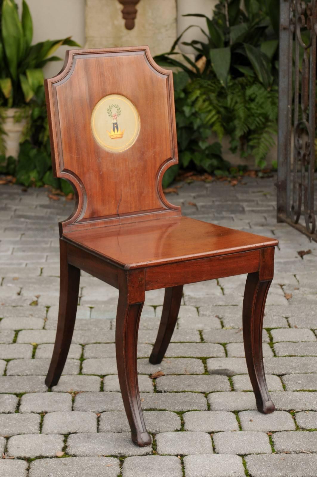 An English wooden hall chair with plank seat and painted crest from the mid-19th century. This English hall chair features a slightly slanted cartouche-shaped back, adorned in its center with medallion depicting a crest made of a crown, from which
